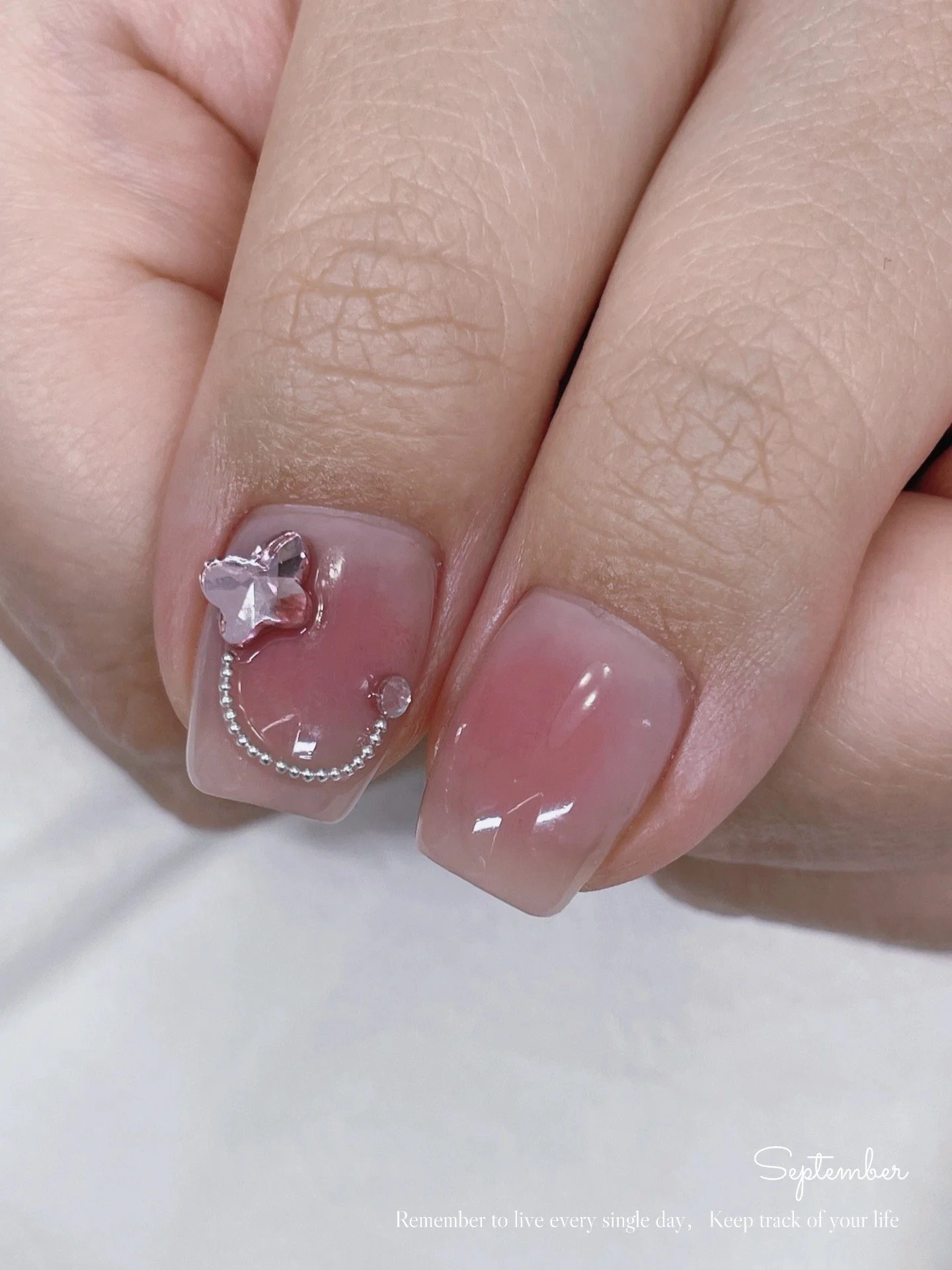Close-up of a hand with a nude pink nail polish on manicured nails, one nail decorated with a silver butterfly-shaped nail charm and small decorative silver beads, against a neutral background.