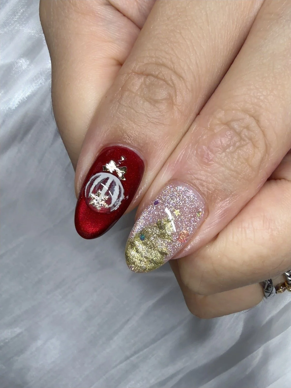 Close-up of hands showing Christmas-themed decorated nails, one with a red base and a white design resembling a Christmas ornament, and the other with glittery gold, silver, and colored dots.