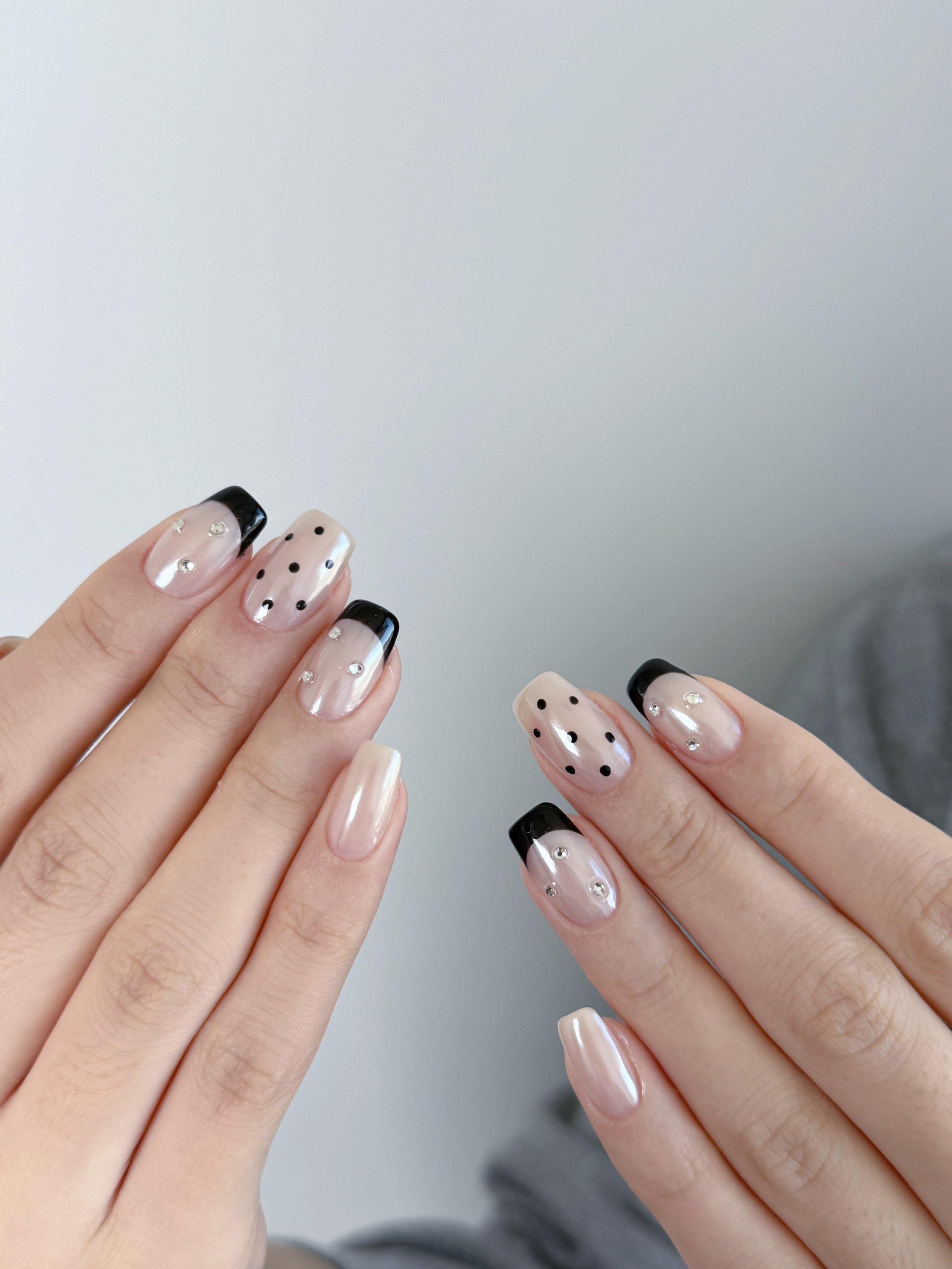 Close-up of hands with manicured nails featuring a French tip with black borders and small rhinestone accents, and nails with black polka dots, on a plain background.
