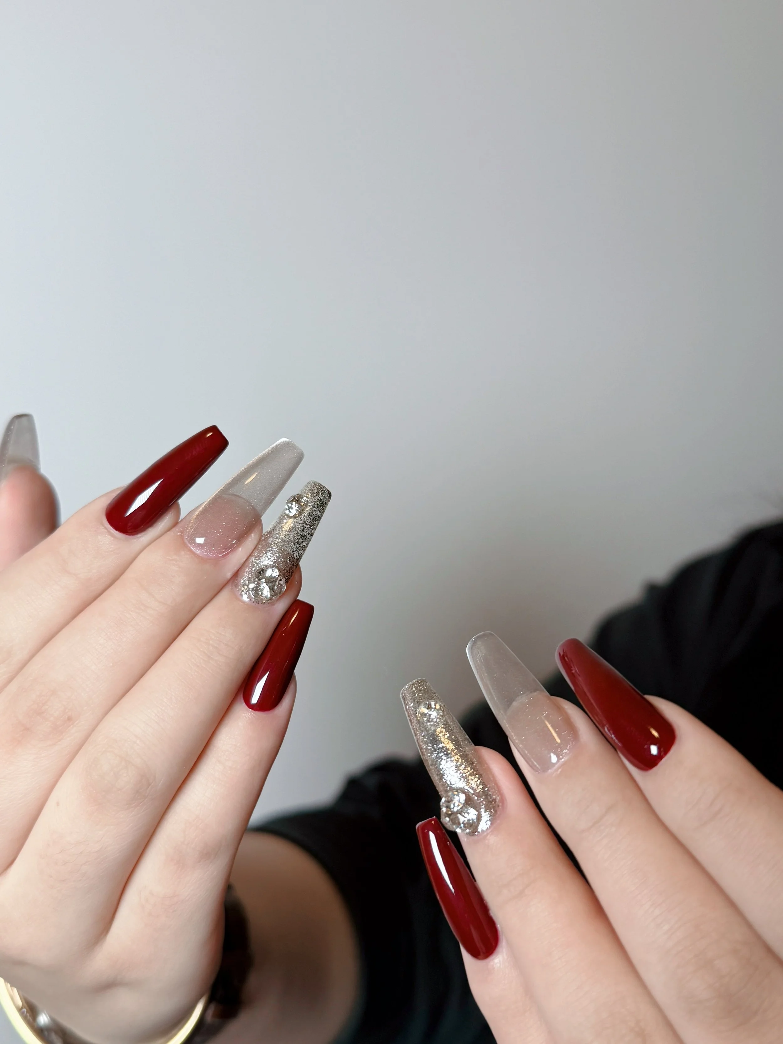 Close-up of hands with manicured nails in red, silver, and glitter designs, some with rhinestones, against a plain background.