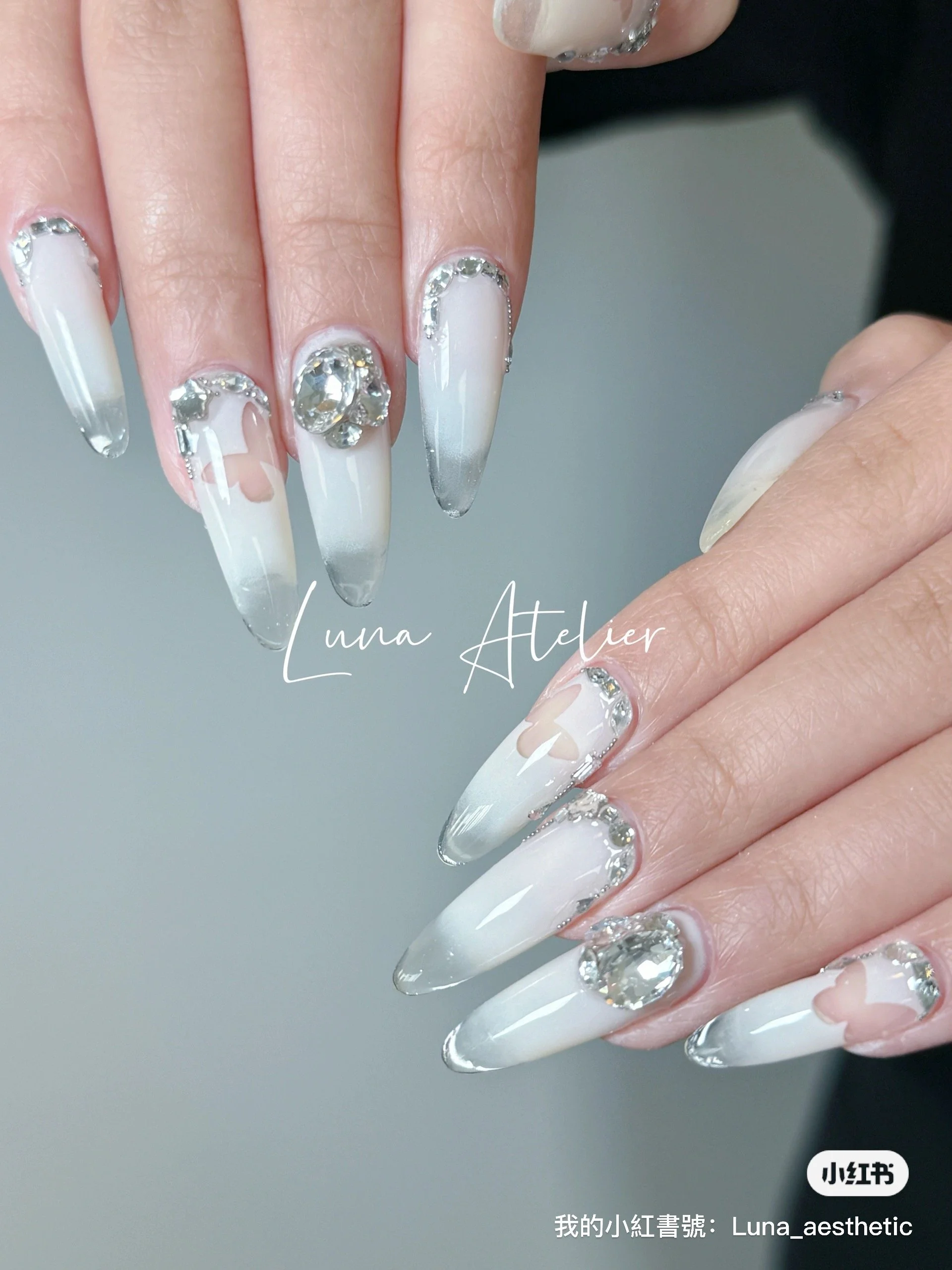Close-up of a hand showing long, almond-shaped nails with white and gray ombre polish, decorated with large rhinestones and heart-shaped accents.