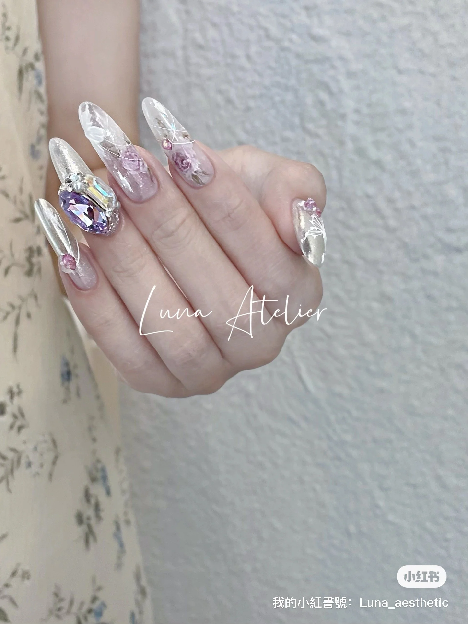 Close-up of a hand with long, decorated acrylic nails featuring a marbled pink and white design, rhinestones, and a large purple gemstone embellishment, set against a textured gray background.
