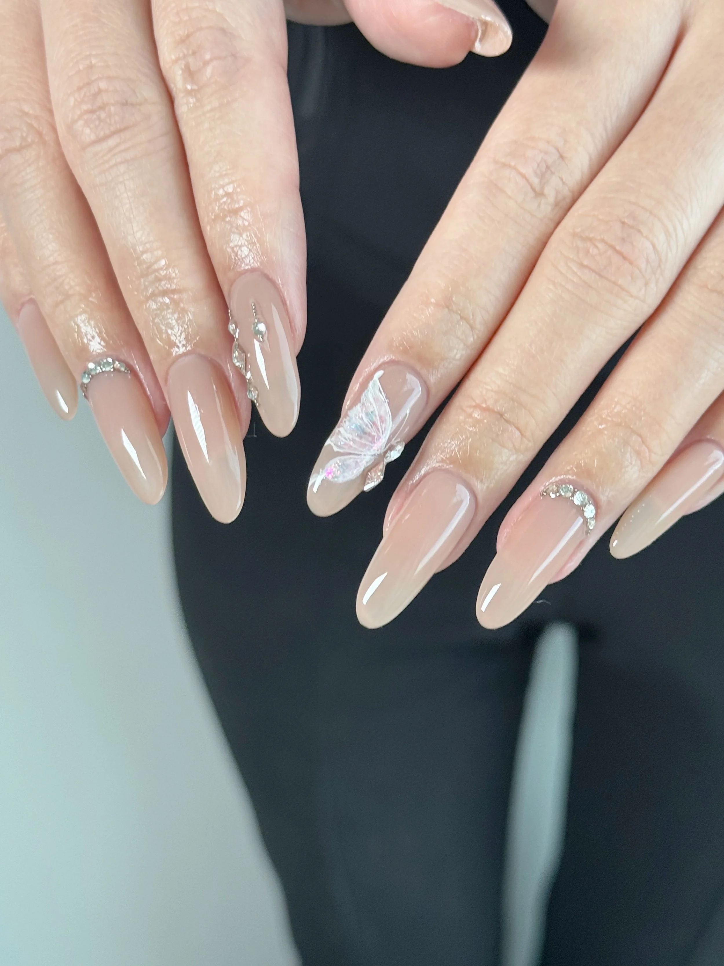 Close-up of manicured hands with nude-colored, elongated oval-shaped nails, decorated with rhinestones and a butterfly design on one nail.