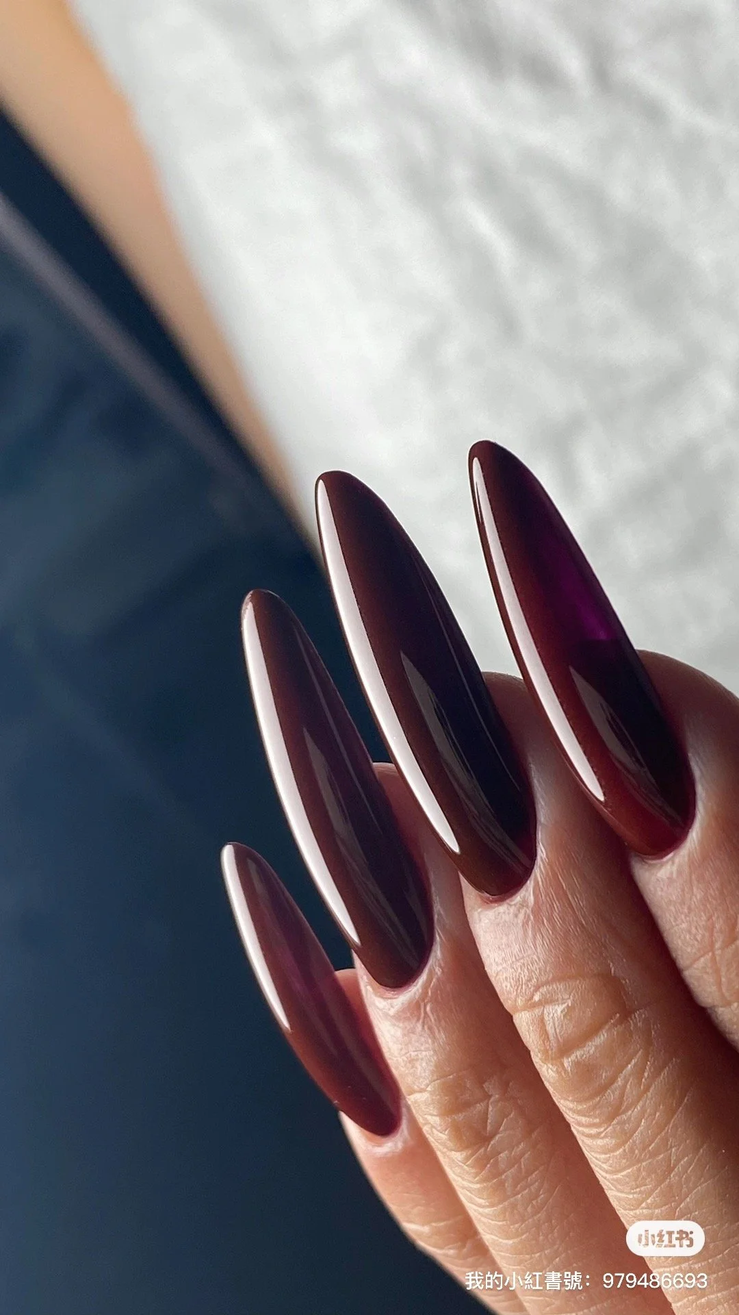 Close-up of a hand with long, almond-shaped dark maroon or black glossy nails. The background is blurred.