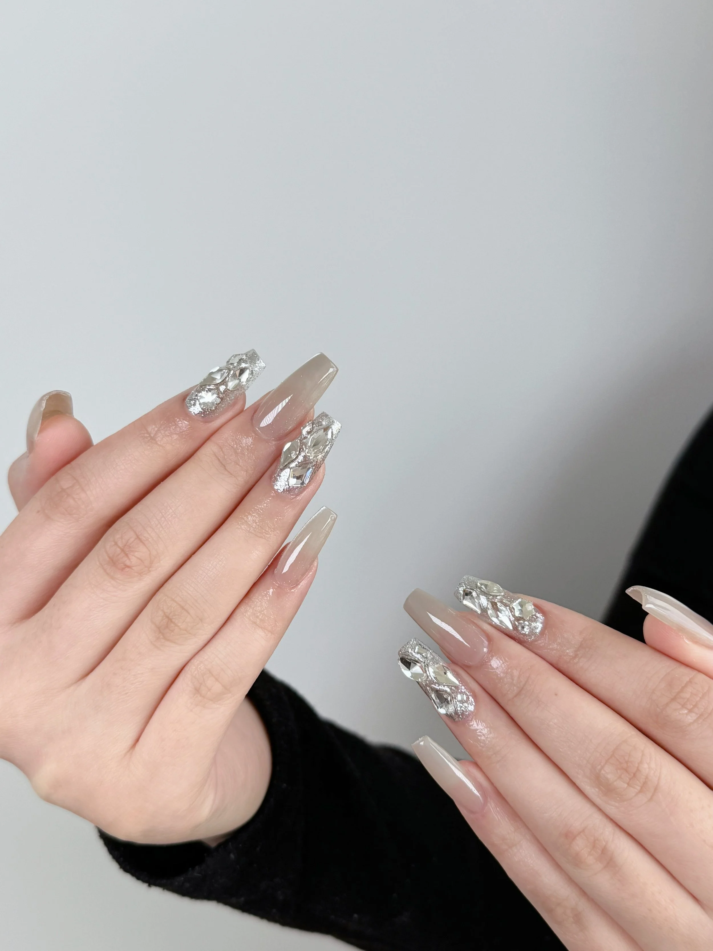 Close-up of hands with long, nude-colored acrylic nails, some featuring silver geometric embellishments, held against a plain background.