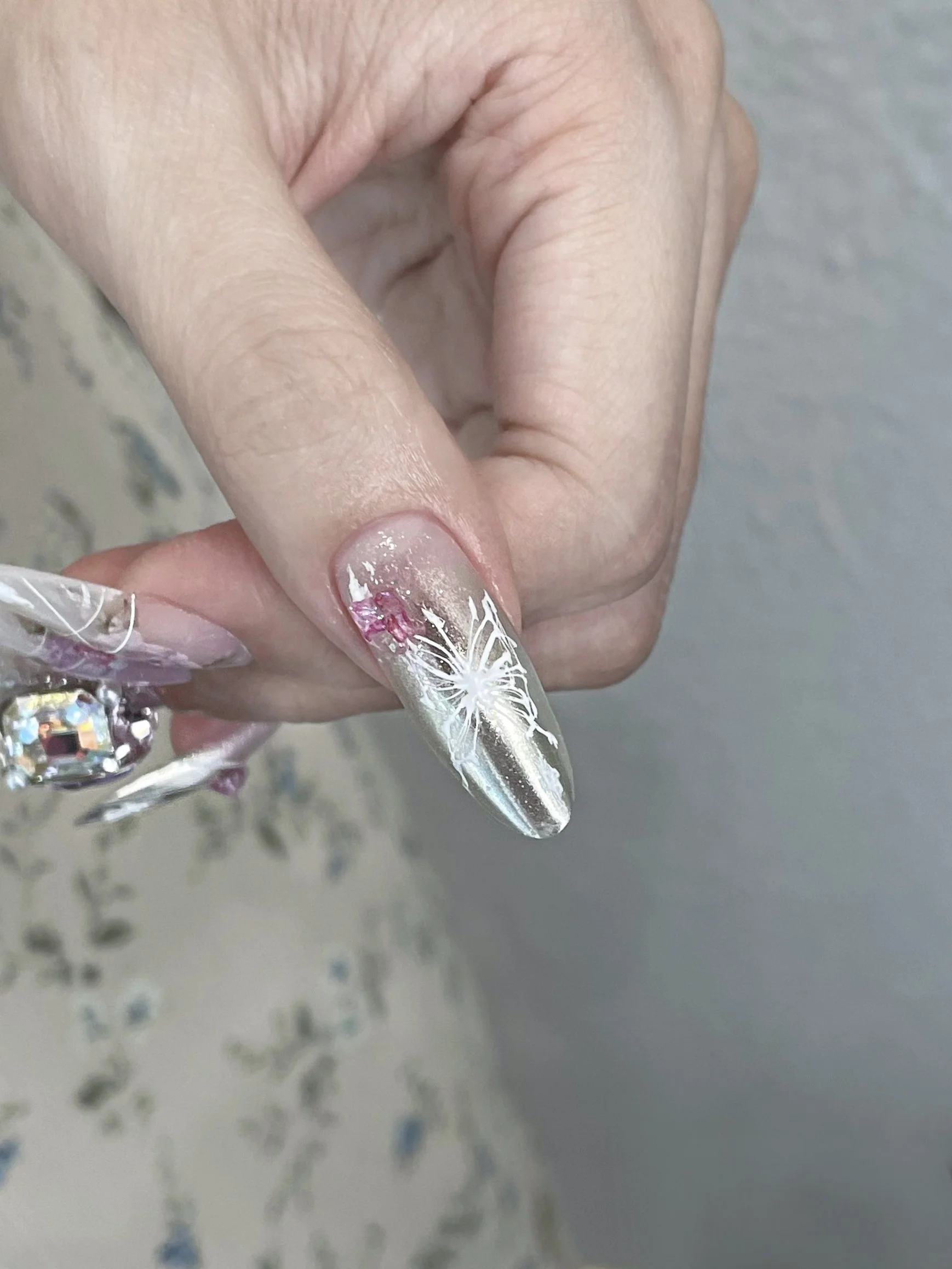 Close-up of a hand with long, metallic silver nail art featuring white floral design on the ring finger.