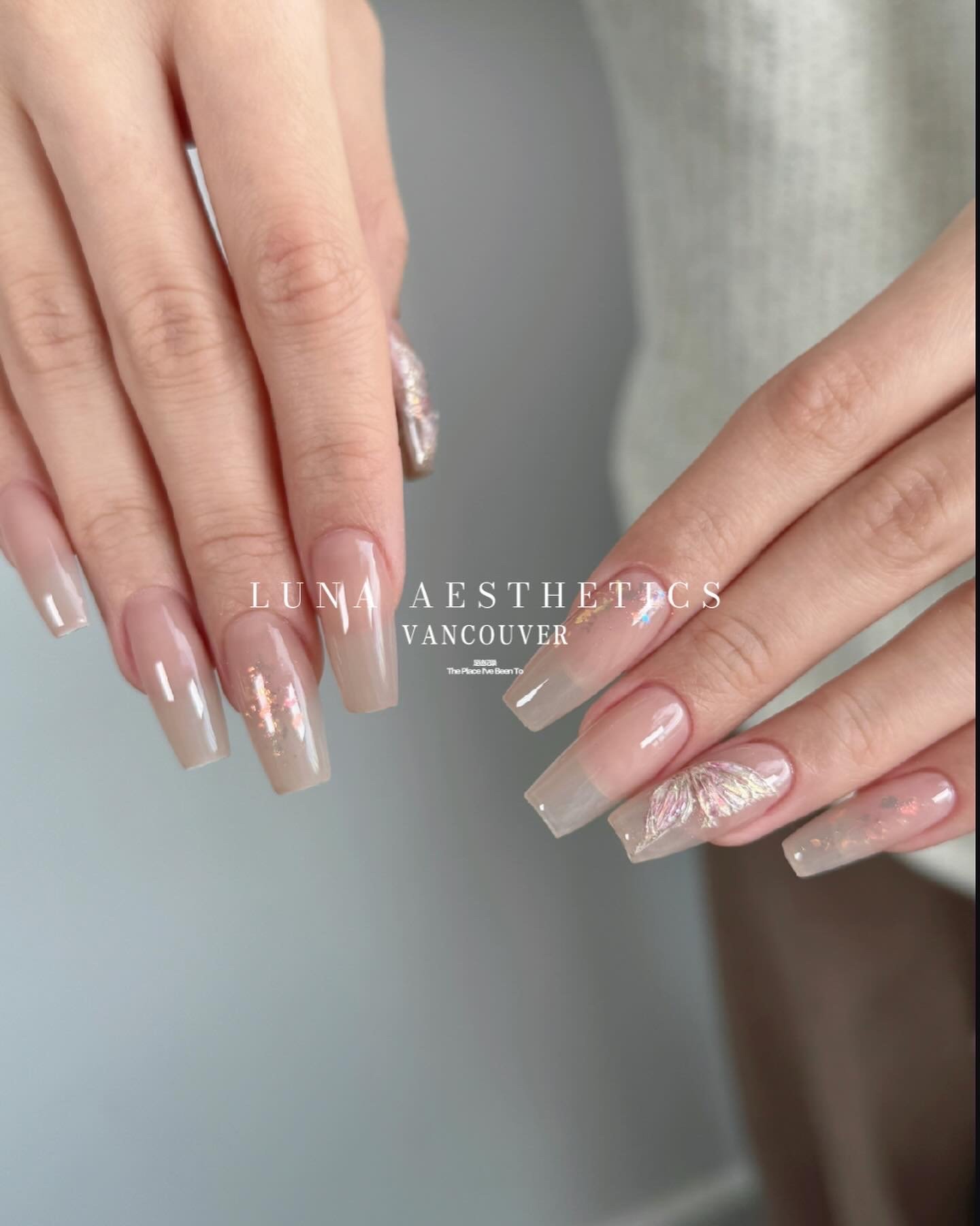 Close-up of a person's hands with manicured nails featuring a light, glossy polish and floral and iridescent accent designs, with the text "LUNA AESTHETICS VANCOUVER" overlayed.