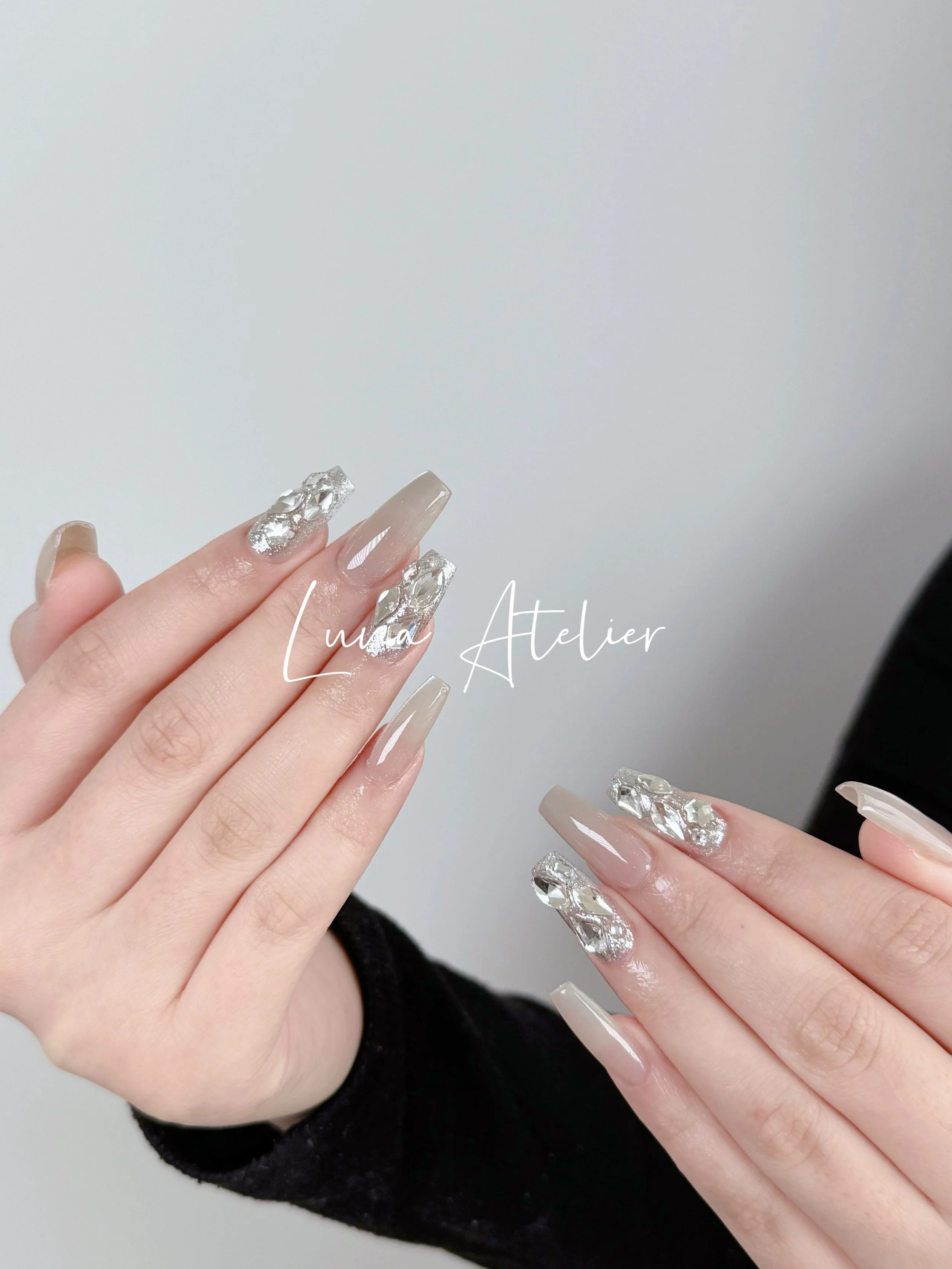 Close-up of hands with long, manicured nails featuring beige and clear polish, with some nails adorned with large, decorative, silver rhinestones. The background is plain and neutral.
