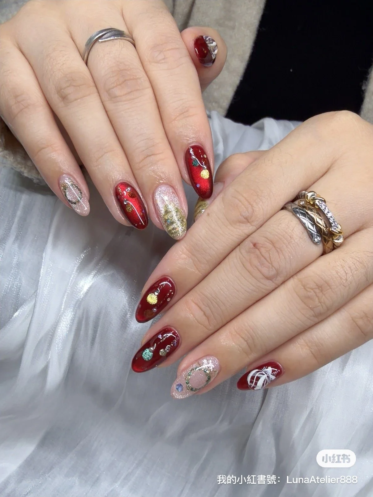 Close-up of a person's hands with decorated nails, some painted in red with gold and multicolored glitter accents, others in a light pinkish shade with gold and glitter designs, and some featuring small embellishments like glitter and decals. The per