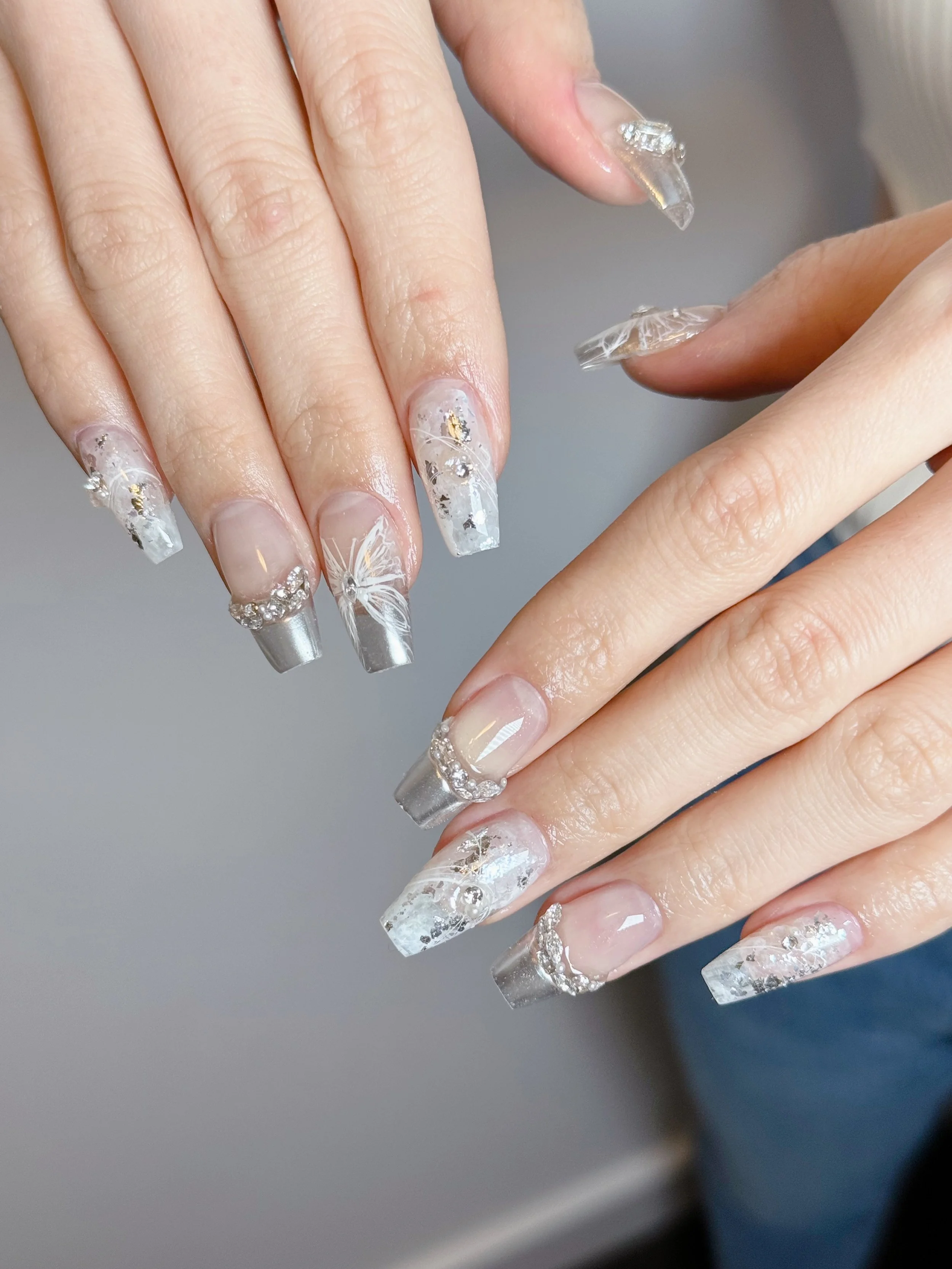 Close-up of hands with long, decorated acrylic nail art featuring clear, silver, and white designs with rhinestones and gold accents.