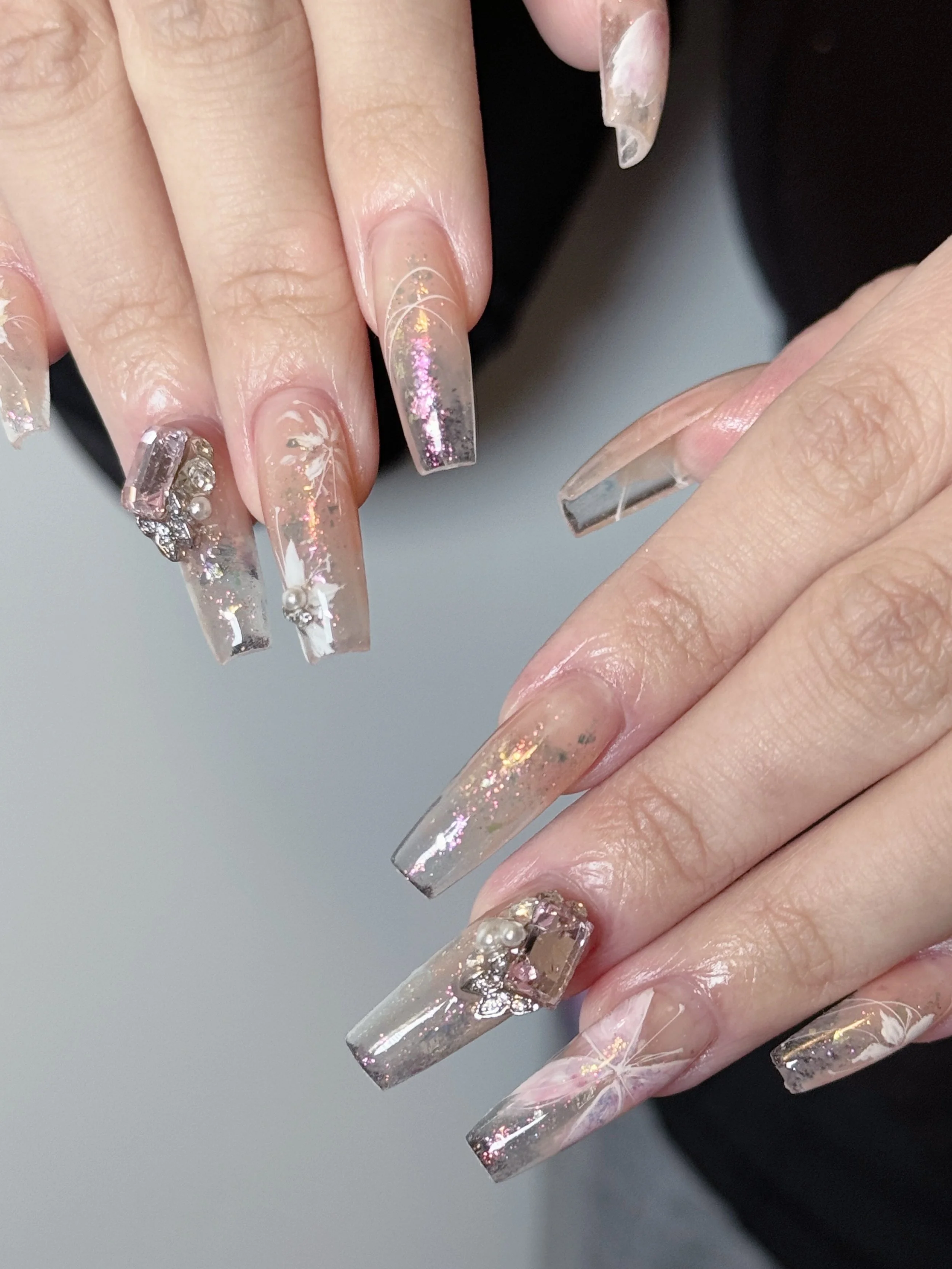 Close-up of manicured nails with long, acrylic extensions, decorated with iridescent glitter, pearls, and floral designs on a natural, light skin tone.