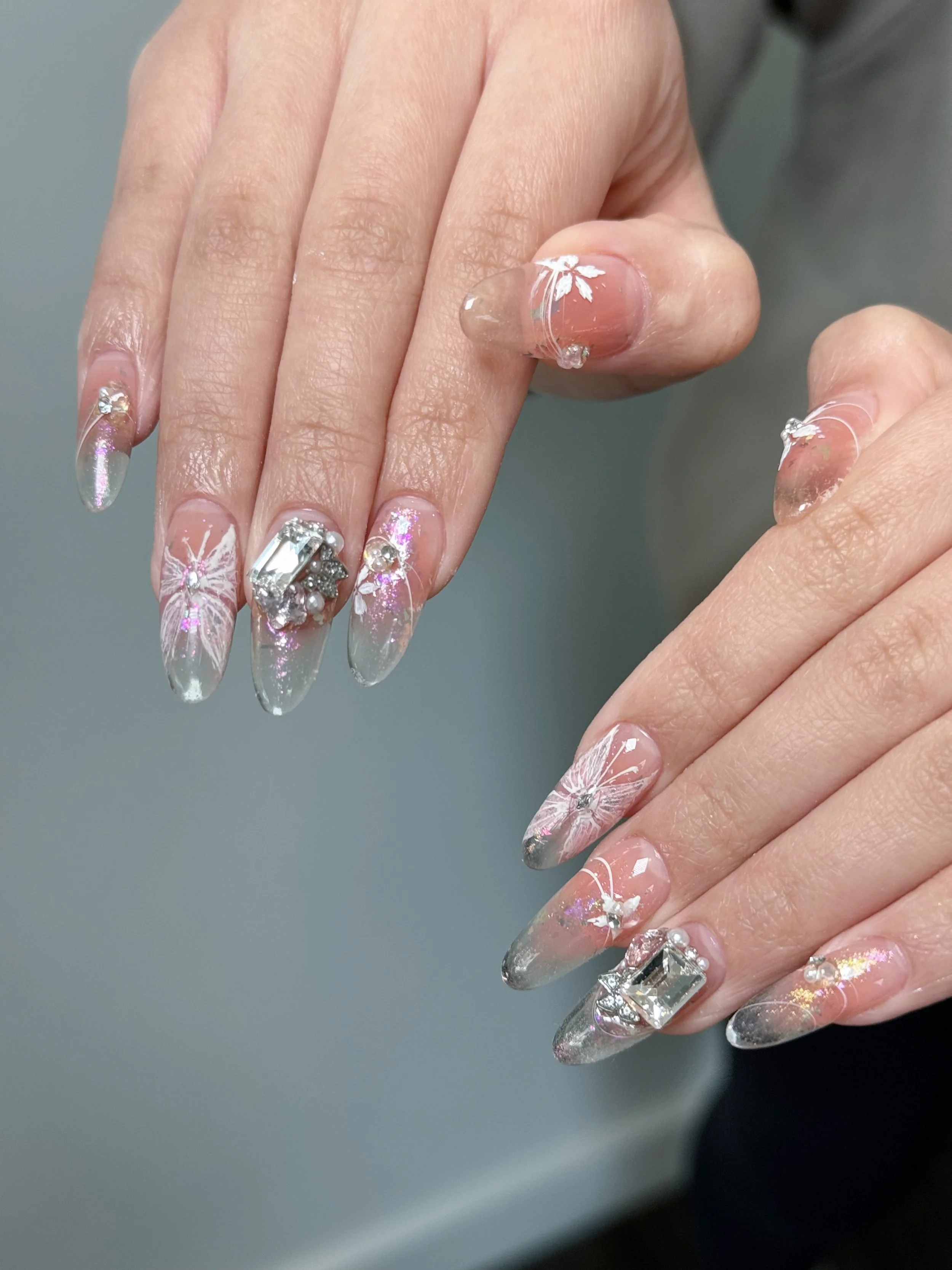 Close-up of manicured nails with glitter, rhinestones, and white floral designs on a clear and pink base