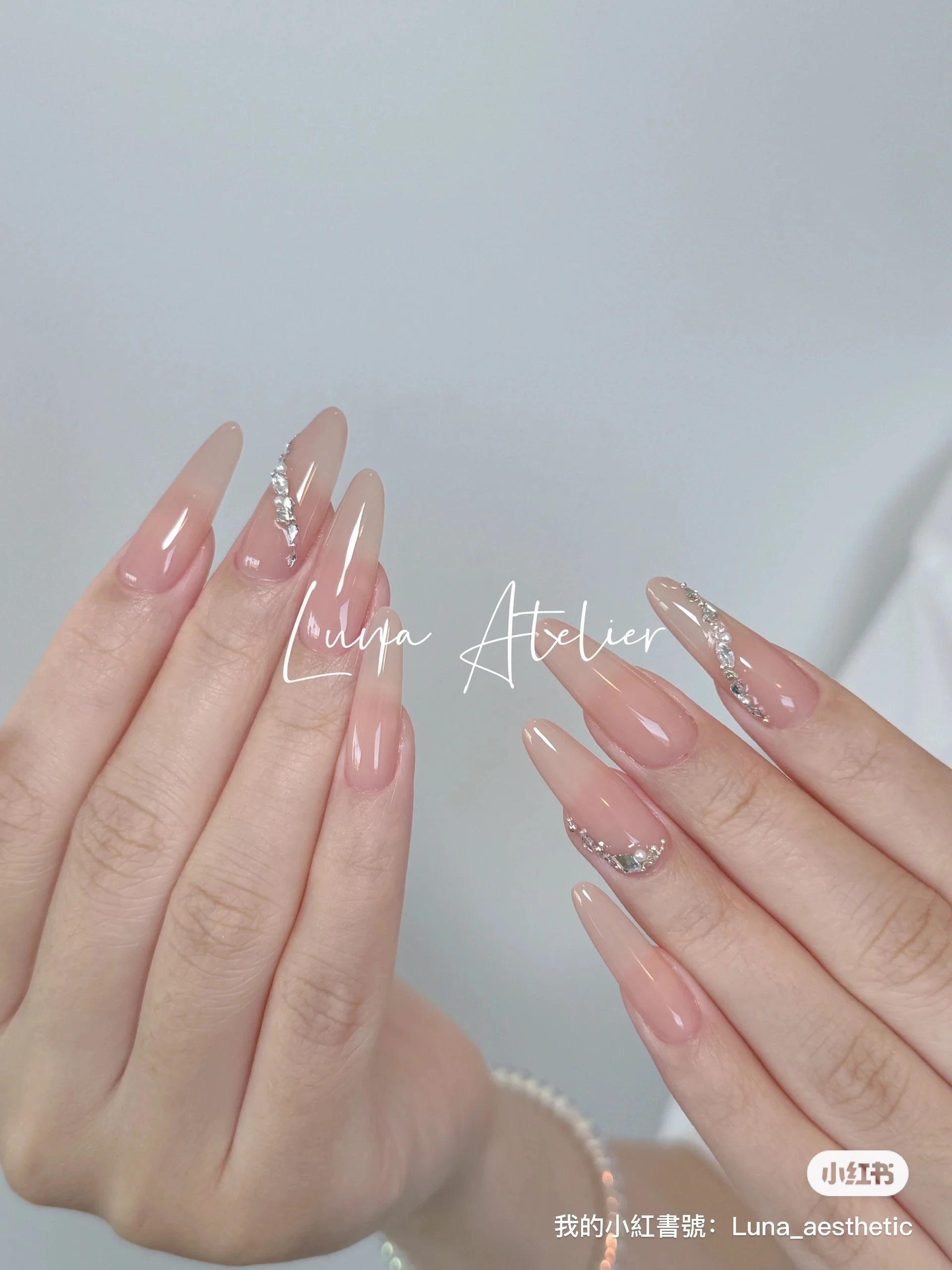 Close-up of manicured nails with a nude pink polish, decorated with small rhinestones on the tips, held against a neutral background.