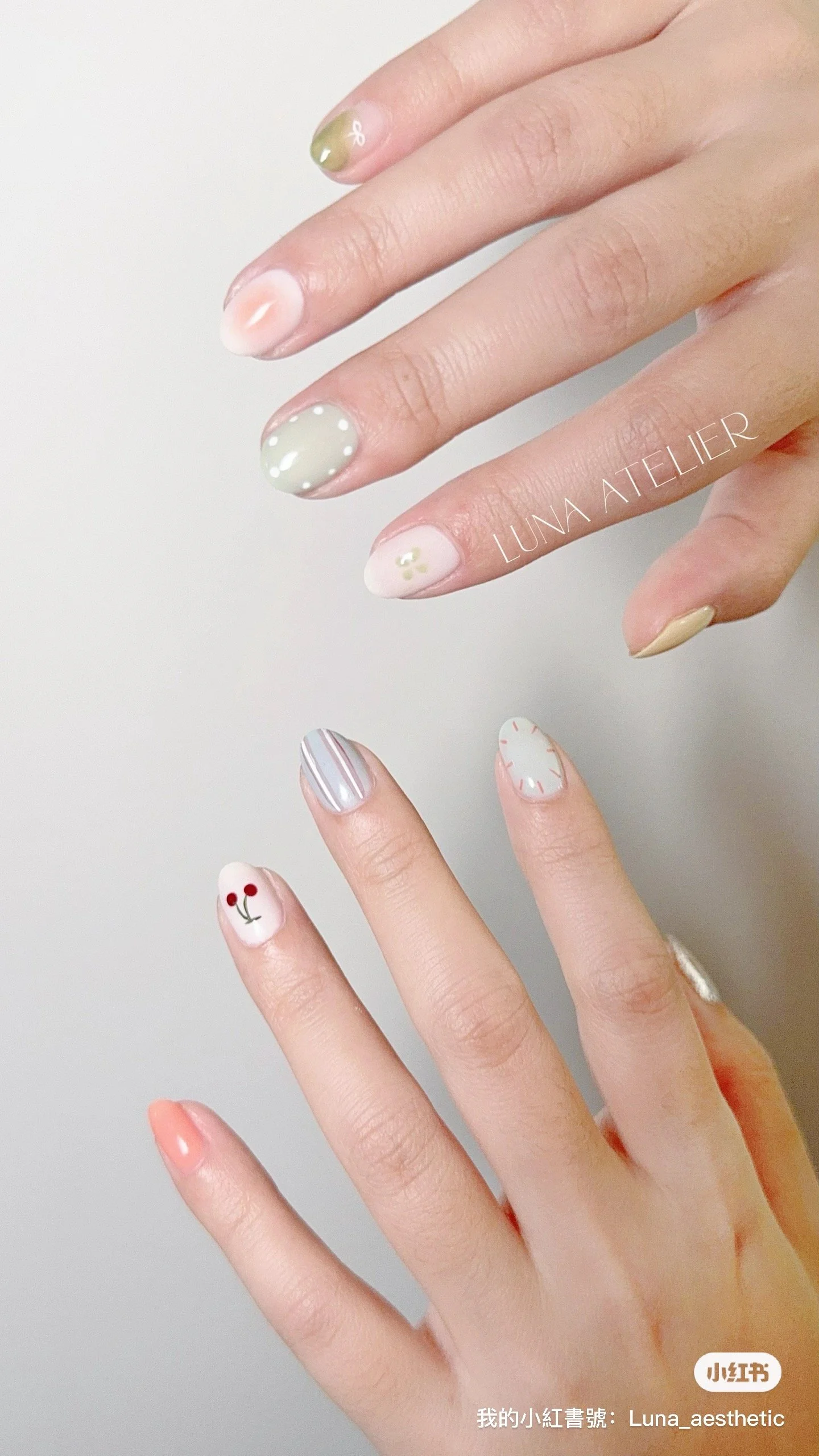 Close-up of two hands with decorated nails. The nails feature various pastel colors and patterns, including polka dots, stripes, a small flower, and cherry designs. The background is plain and light-colored.