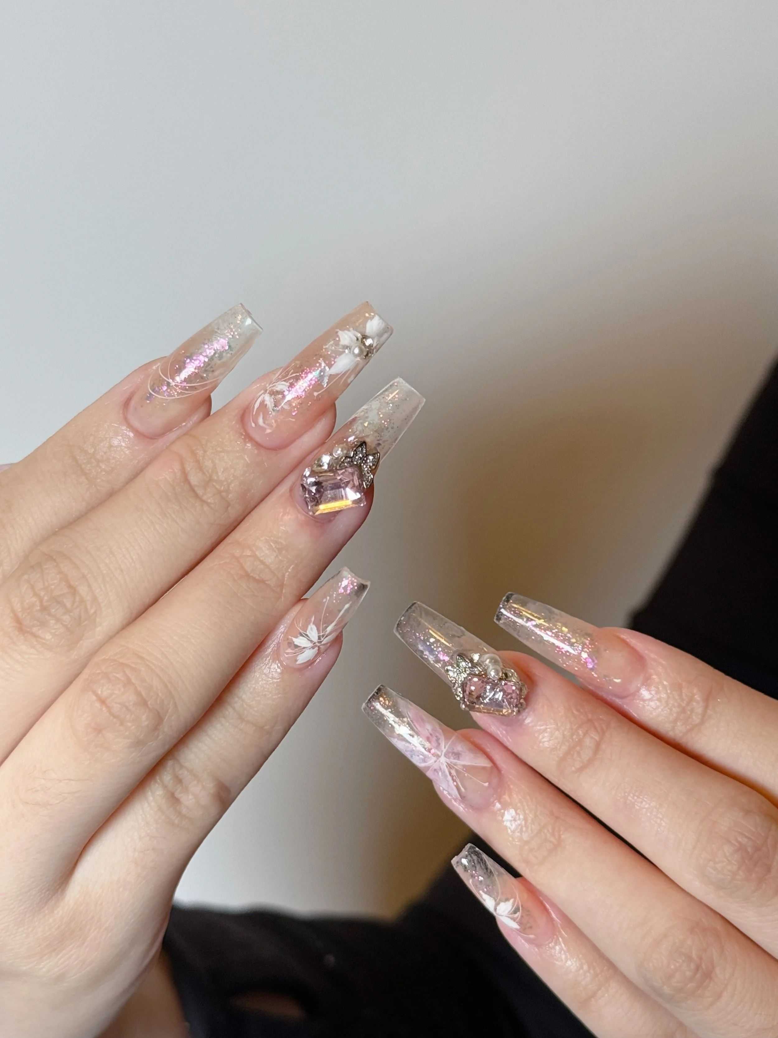 Close-up of decorated acrylic nails with pink and white floral designs, pearls, rhinestones, and glitter on a person's hands.