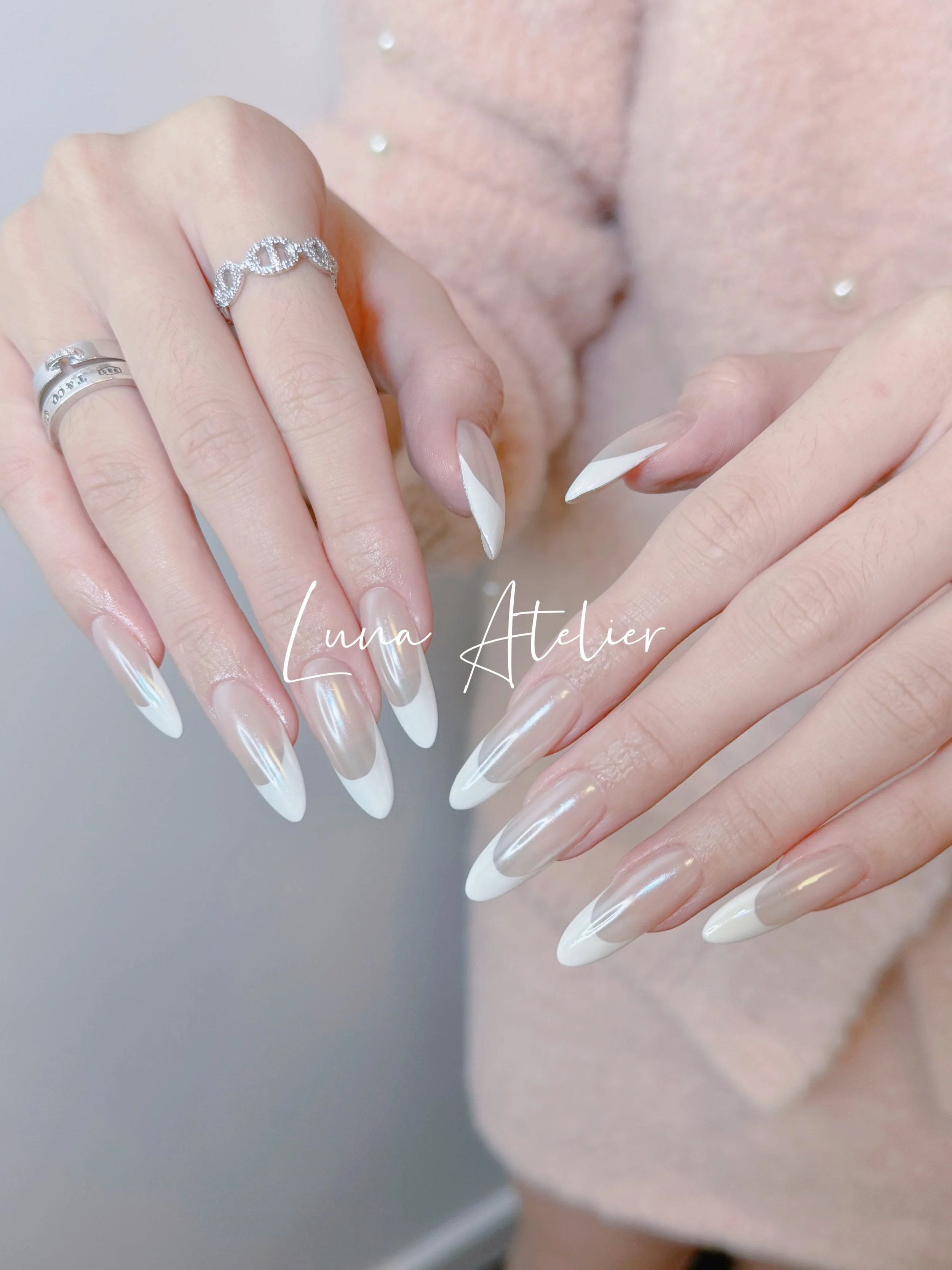Close-up of a woman's hands with professionally manicured nails featuring a white French tip design. She is wearing multiple rings, including a silver band and a ring with 'Love' engraved, and is dressed in a pink textured garment.