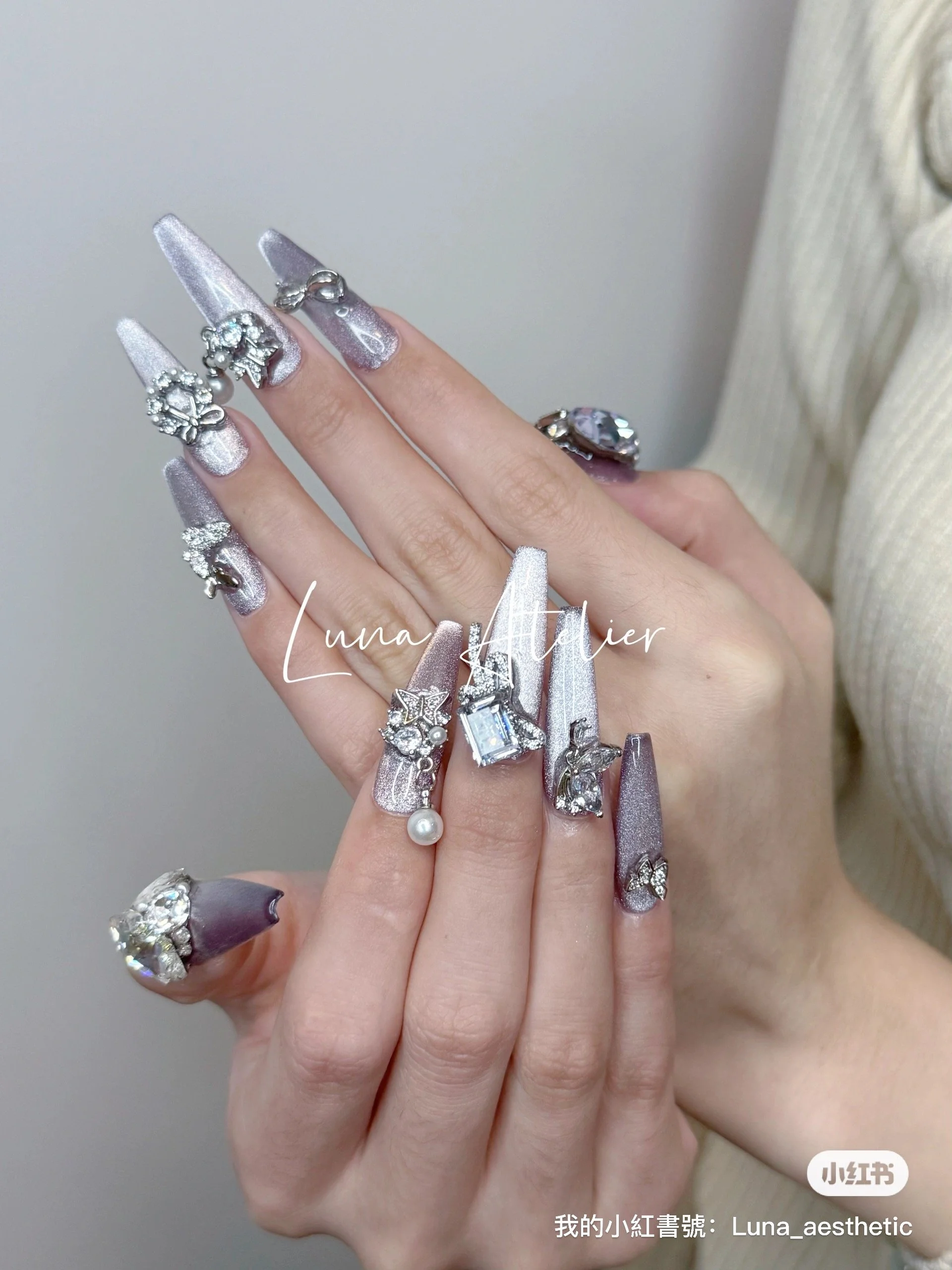 Hand with long, manicured nails painted in light purple with glitter, wearing multiple silver and rhinestone rings, some with pearls or large gemstones, against a plain background.