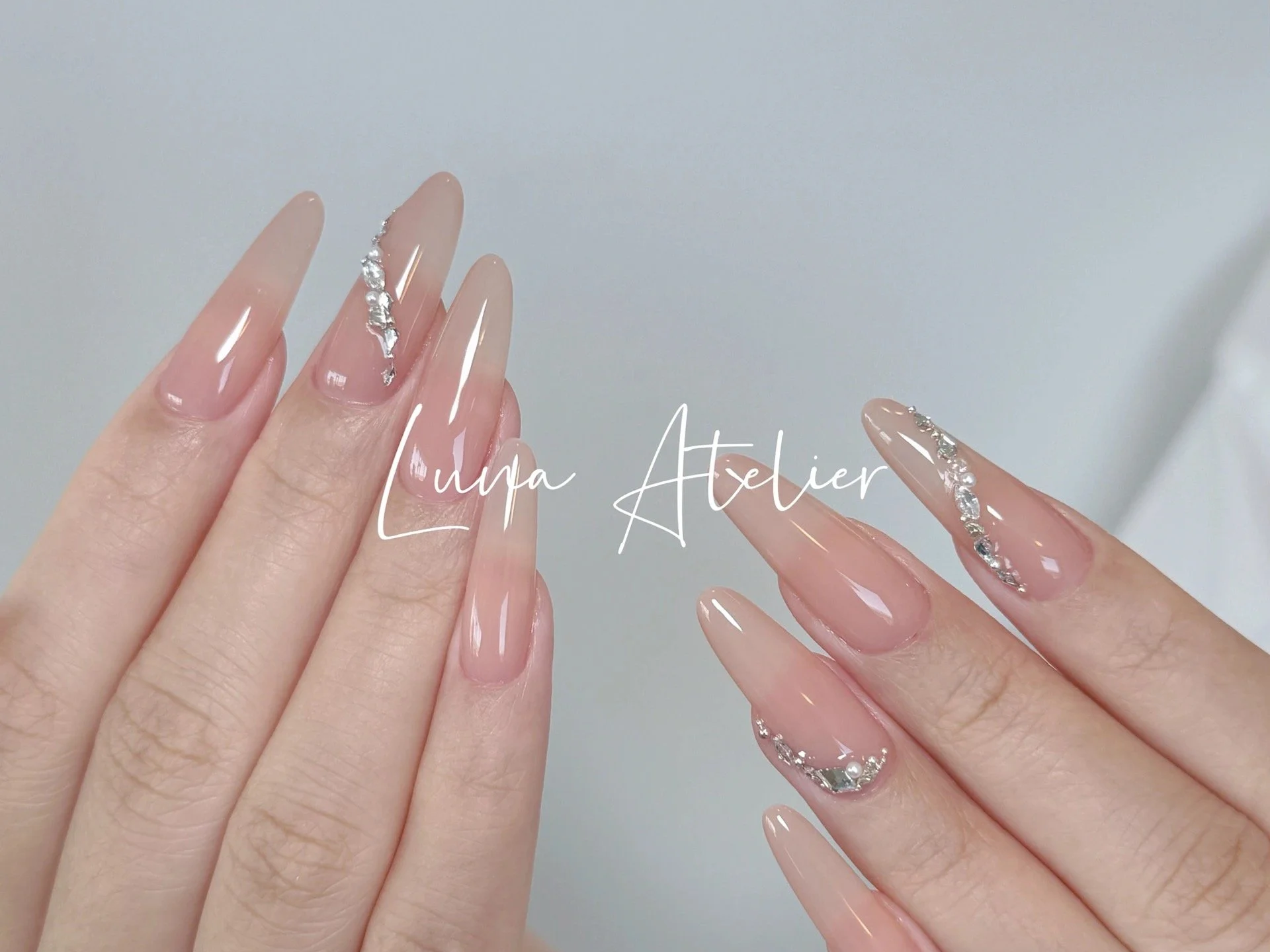 Close-up of two hands with long, almond-shaped nails painted in a nude color, each decorated with silver rhinestones and crystals along the tips.