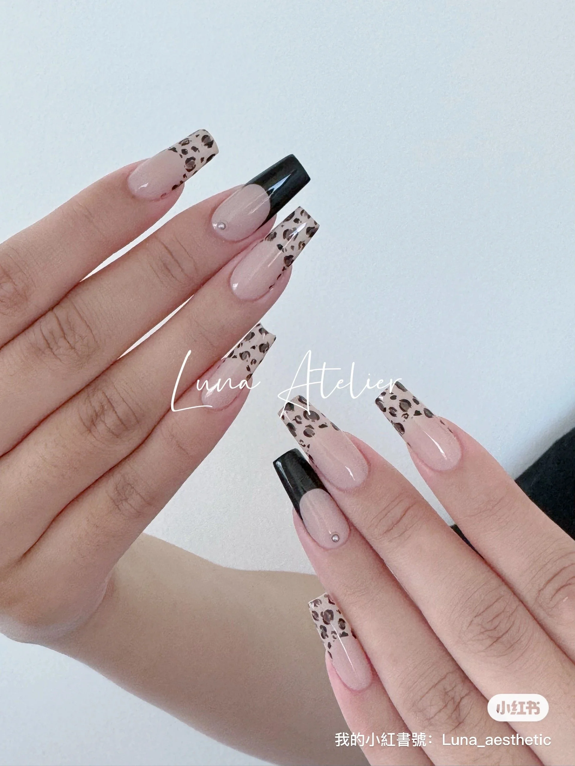 Close-up of hands with manicured nails, featuring a leopard print design on some nails and black French tips, against a plain white background.