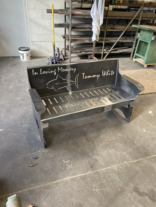 Black metal bench with a chalk drawing of a fish and the words "In Loving Memory Tammy White" written on the backrest, in a workshop or warehouse setting.