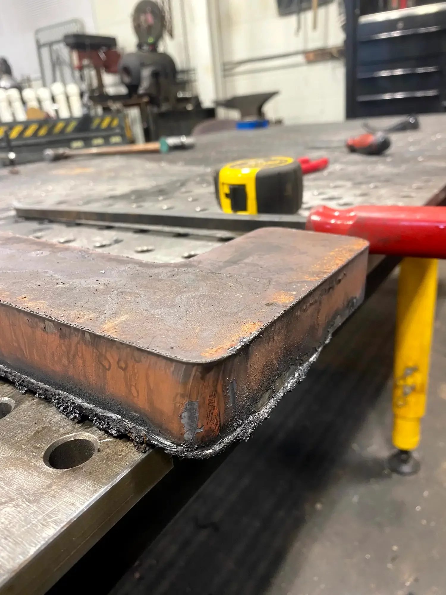 Close-up of a metal workbench with tools, including a yellow tape measure, a red tool, and a welding clamp, in a workshop setting.