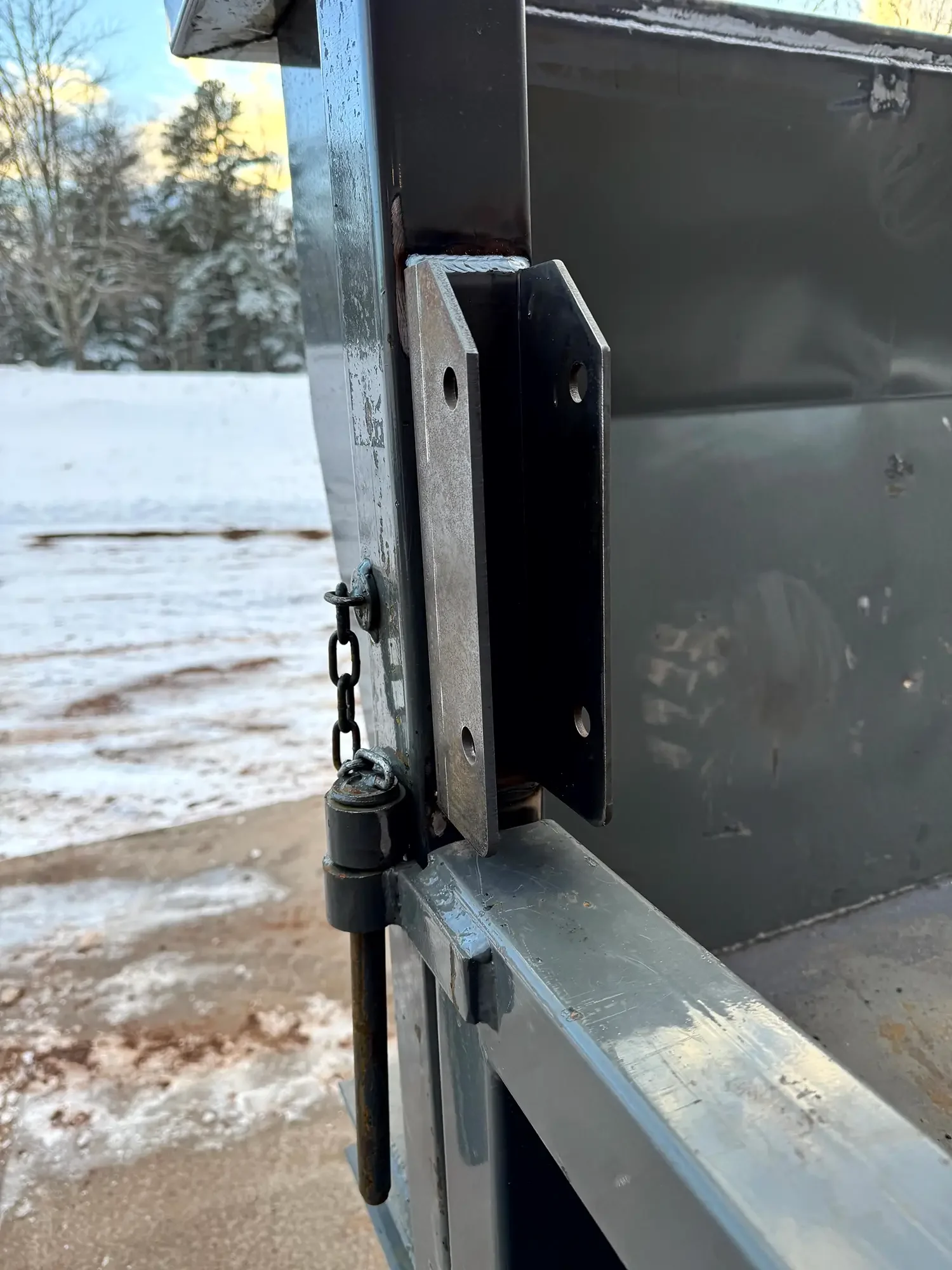Close-up of a trailer hitch with a chain, latch, and metal frame, against a snowy outdoor background with trees.