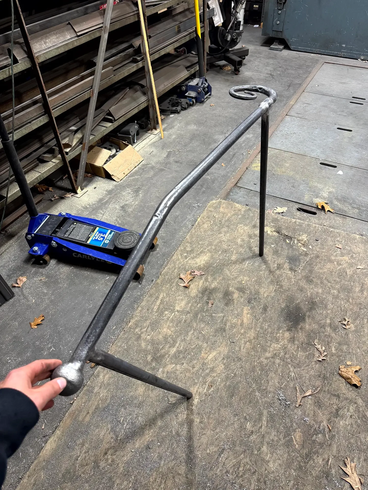 A person's hand holding a metal bar inside a workshop or factory with shelves, tools, and equipment visible on the background floor.