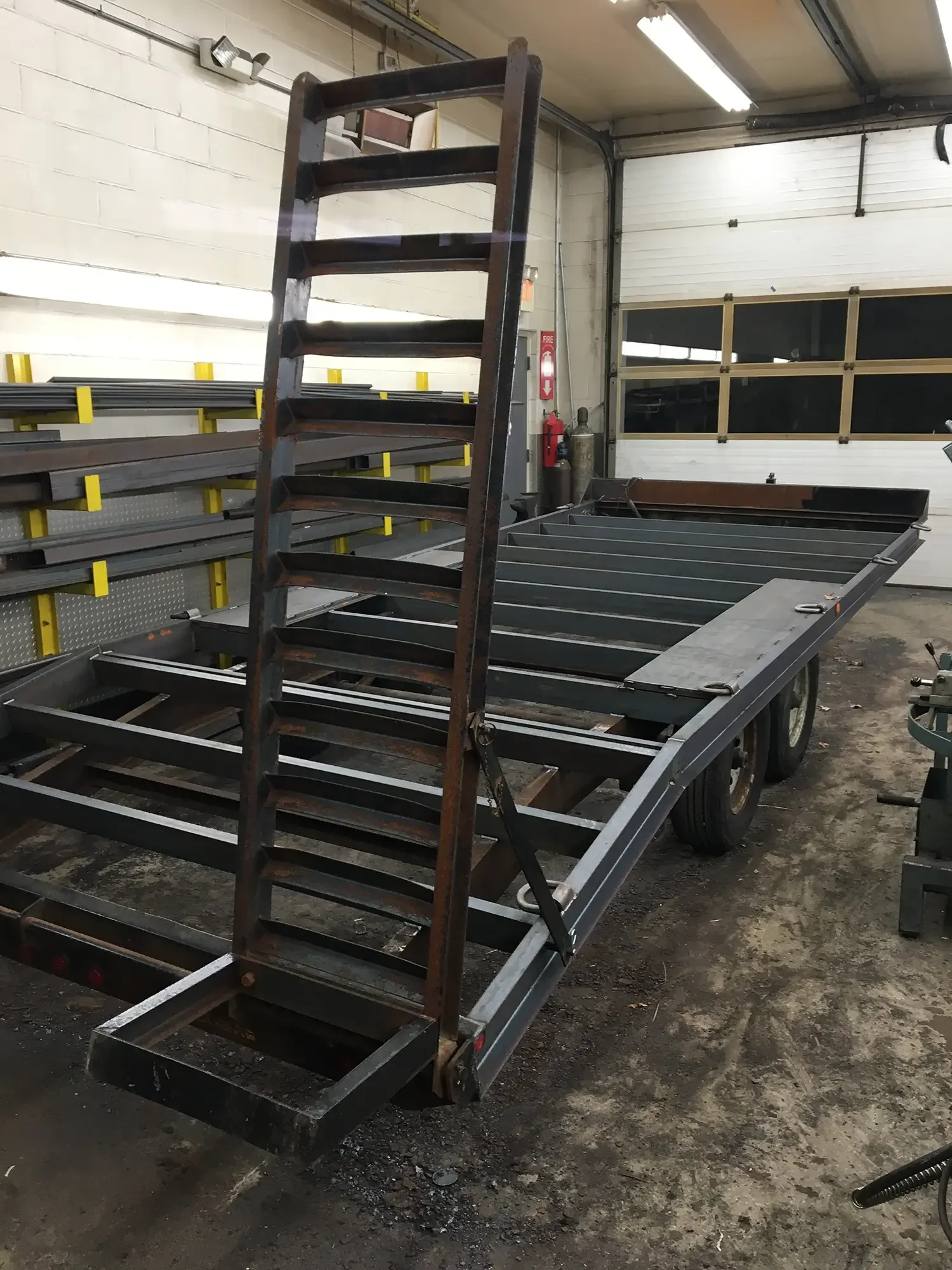 An industrial workshop with a metal frame being assembled on a trailer, with metal shelf racks and equipment around.