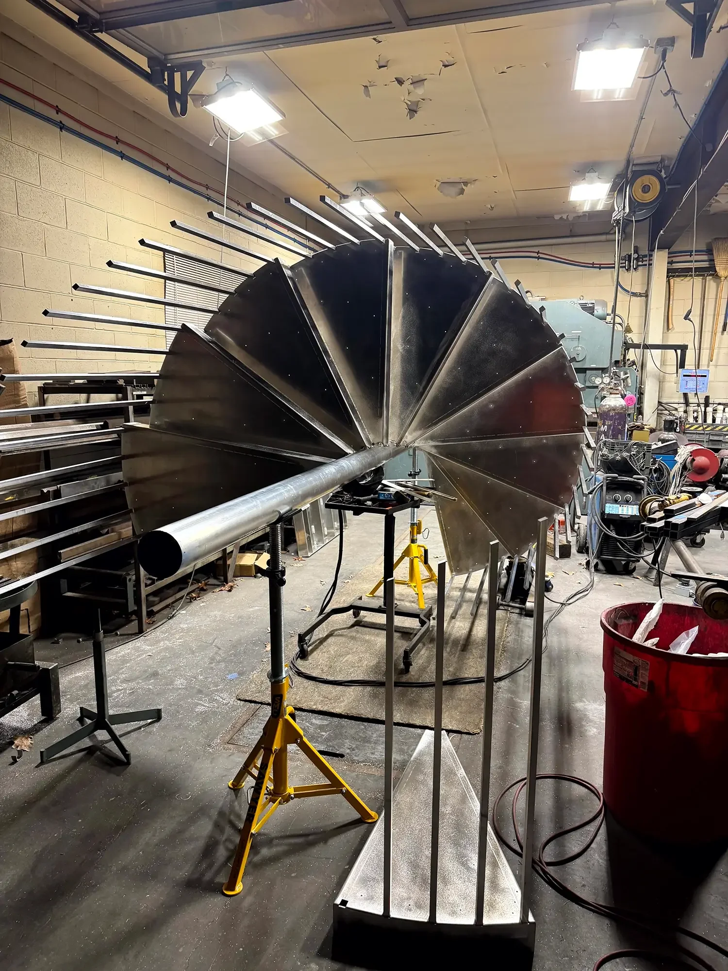 A metallic turbine or propeller with multiple blades is being assembled indoors. The workshop has tools, equipment, and supplies scattered around, with a work table and yellow supports holding the turbine in place. There are bright ceiling lights and