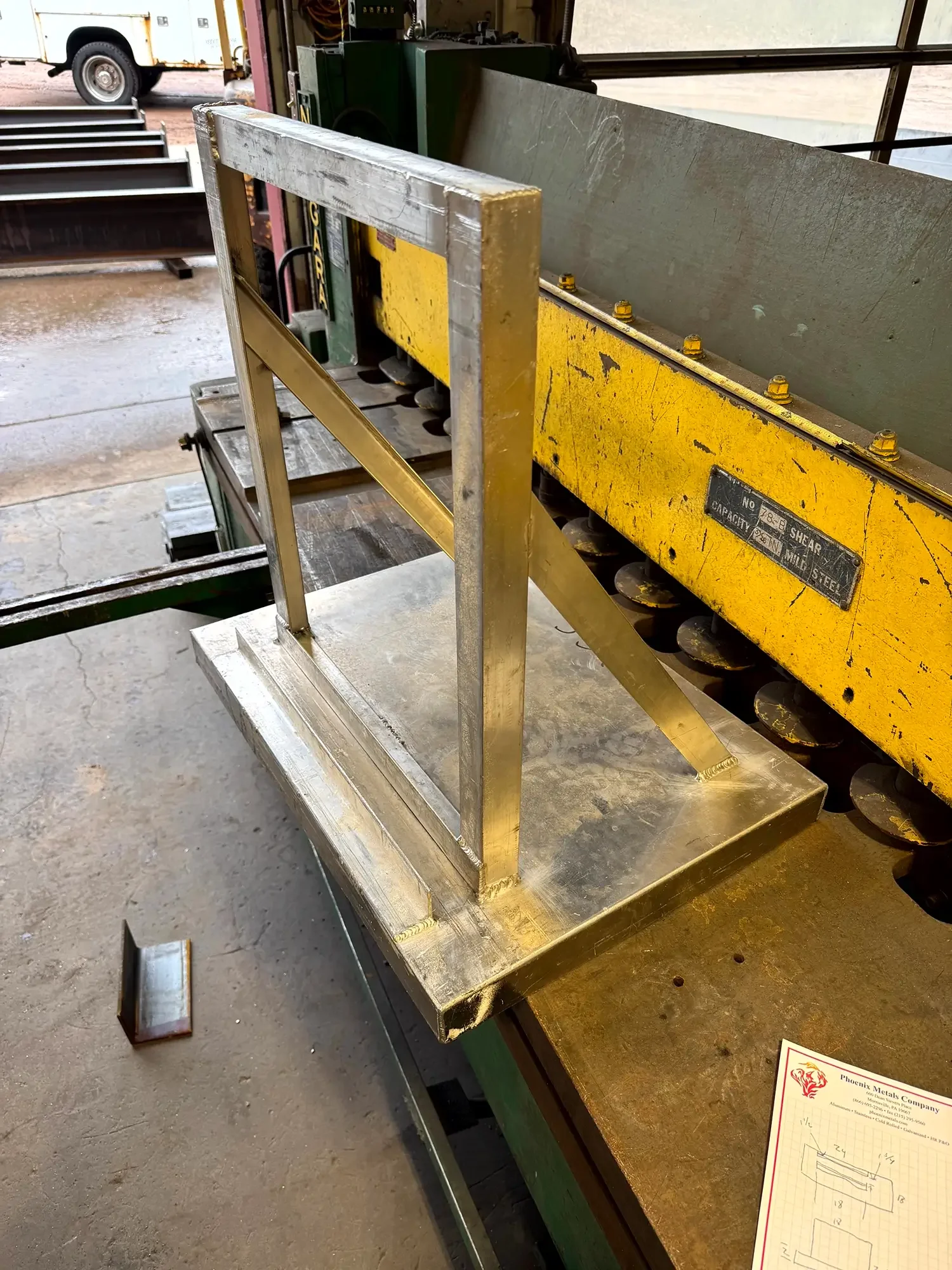 A metal worktable with a metal frame attached, situated in a workshop or industrial setting. In the background, there is a yellow and black machine, and outside, a white utility truck is visible through an open garage door.