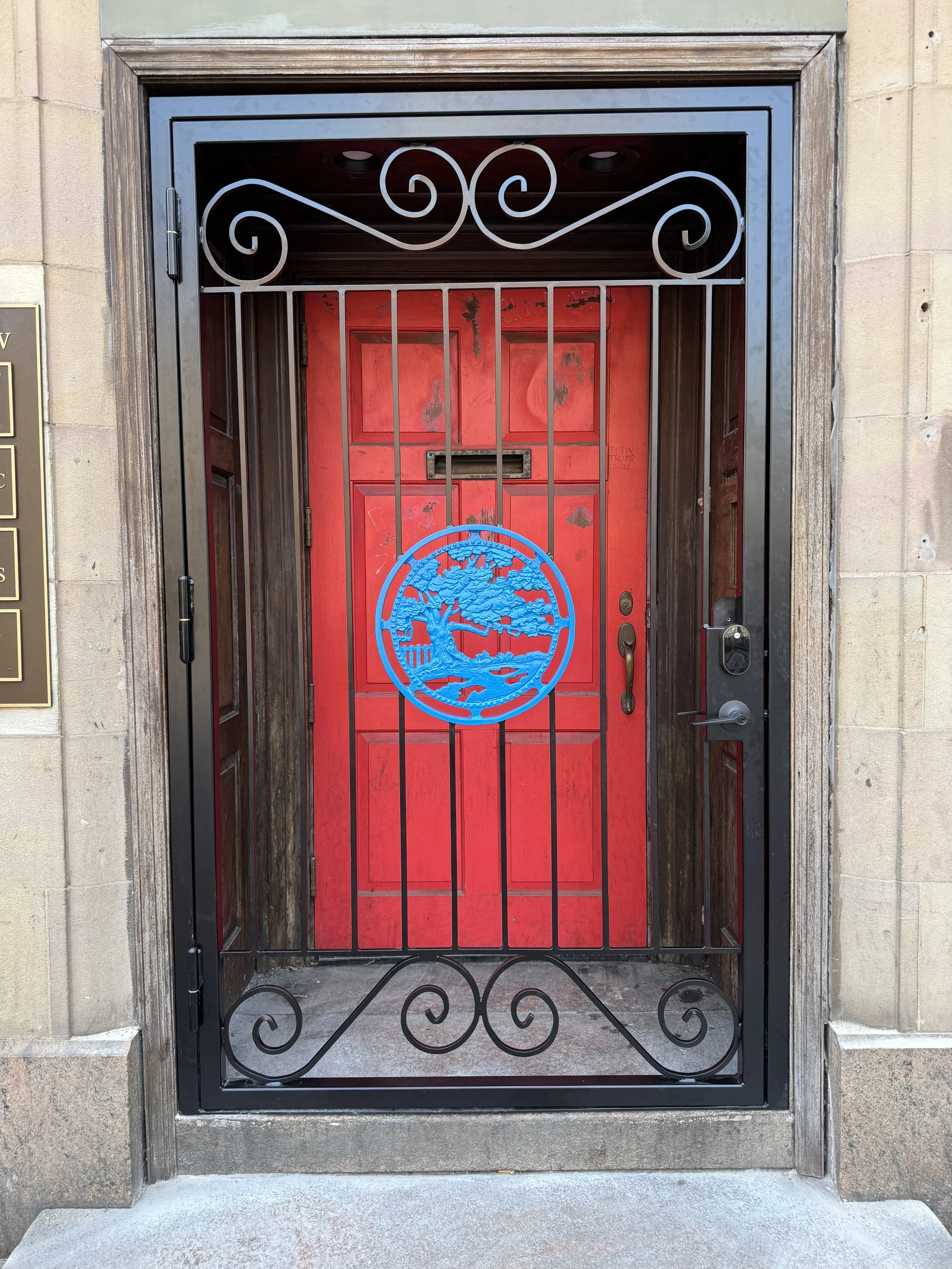 Red front door with a mail slot, behind a black wrought iron security gate with a blue circular decorative emblem of a tree and landscape; the door is set in a stone building with a wooden frame.