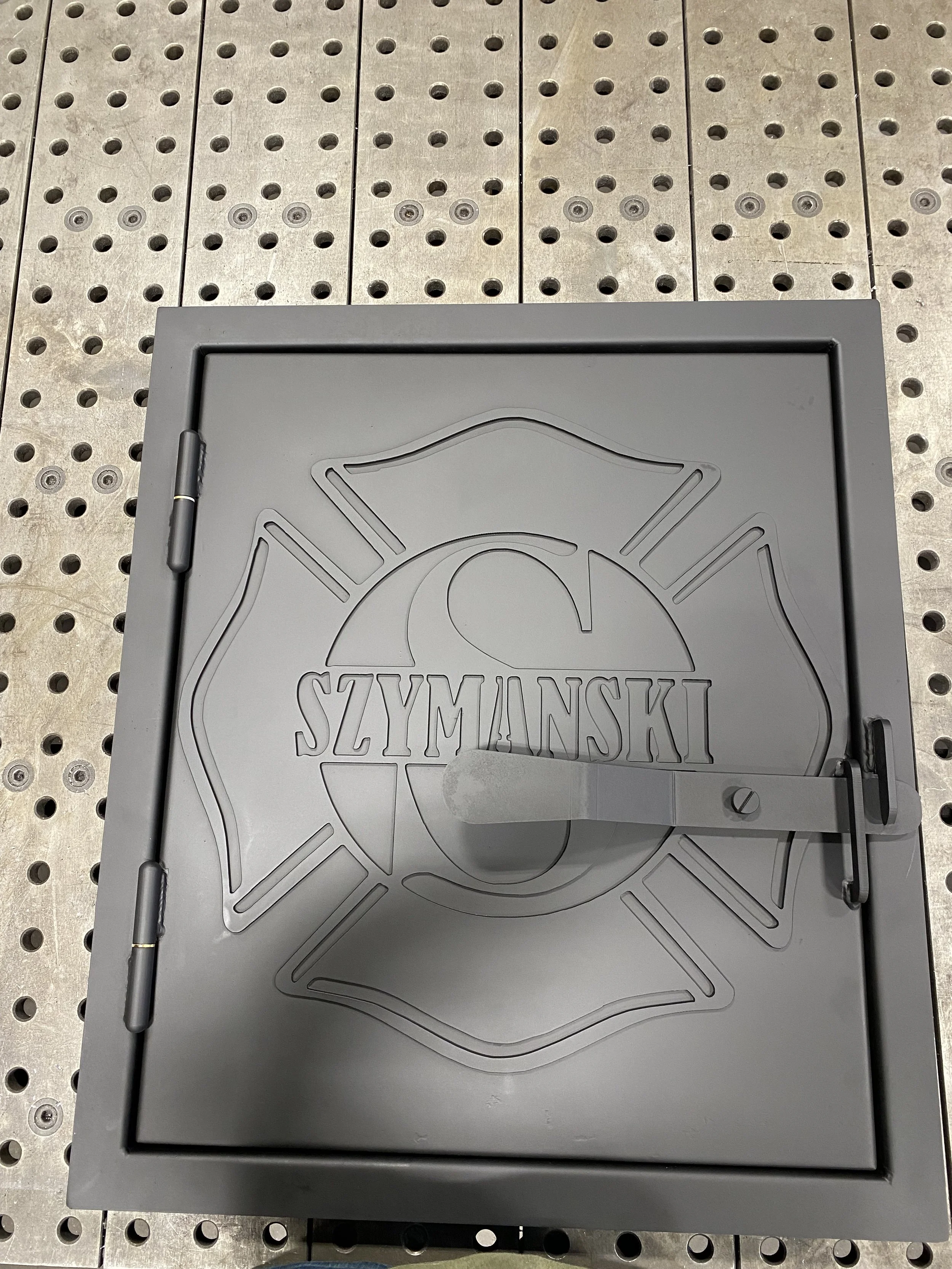 Gray metal fire door with a firefighter emblem and the word 'SZYMANDSKI' embossed on it, placed on a perforated metal floor.