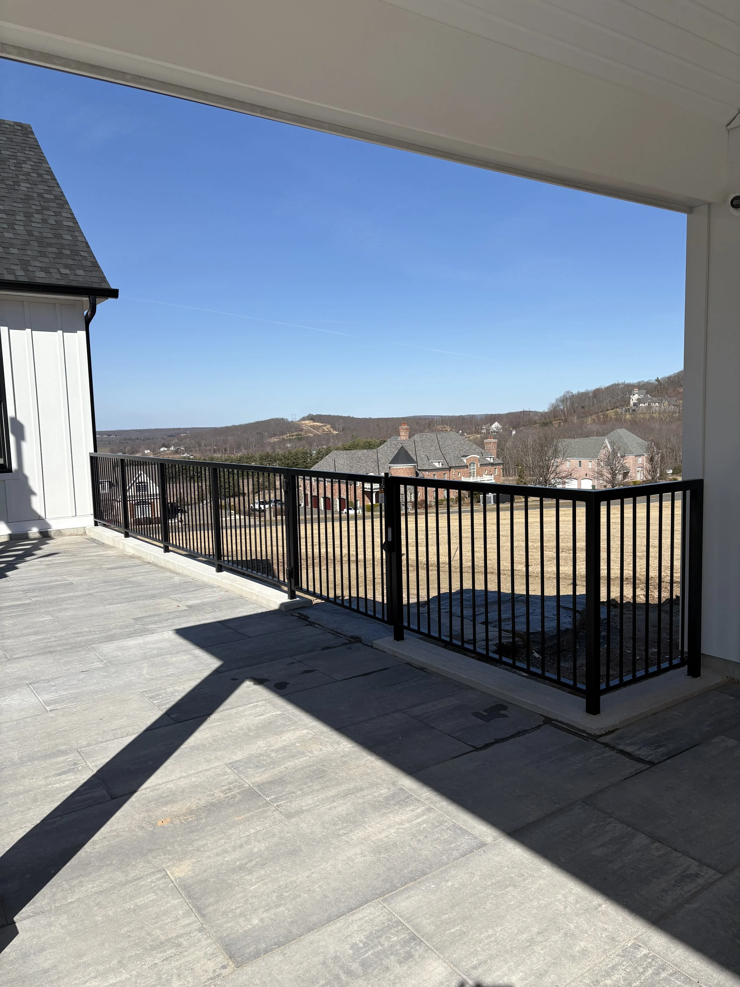 View from a porch or balcony showing a black metal railing, gray tiled floor, and a distant landscape of houses, fields, and hills under a clear blue sky.