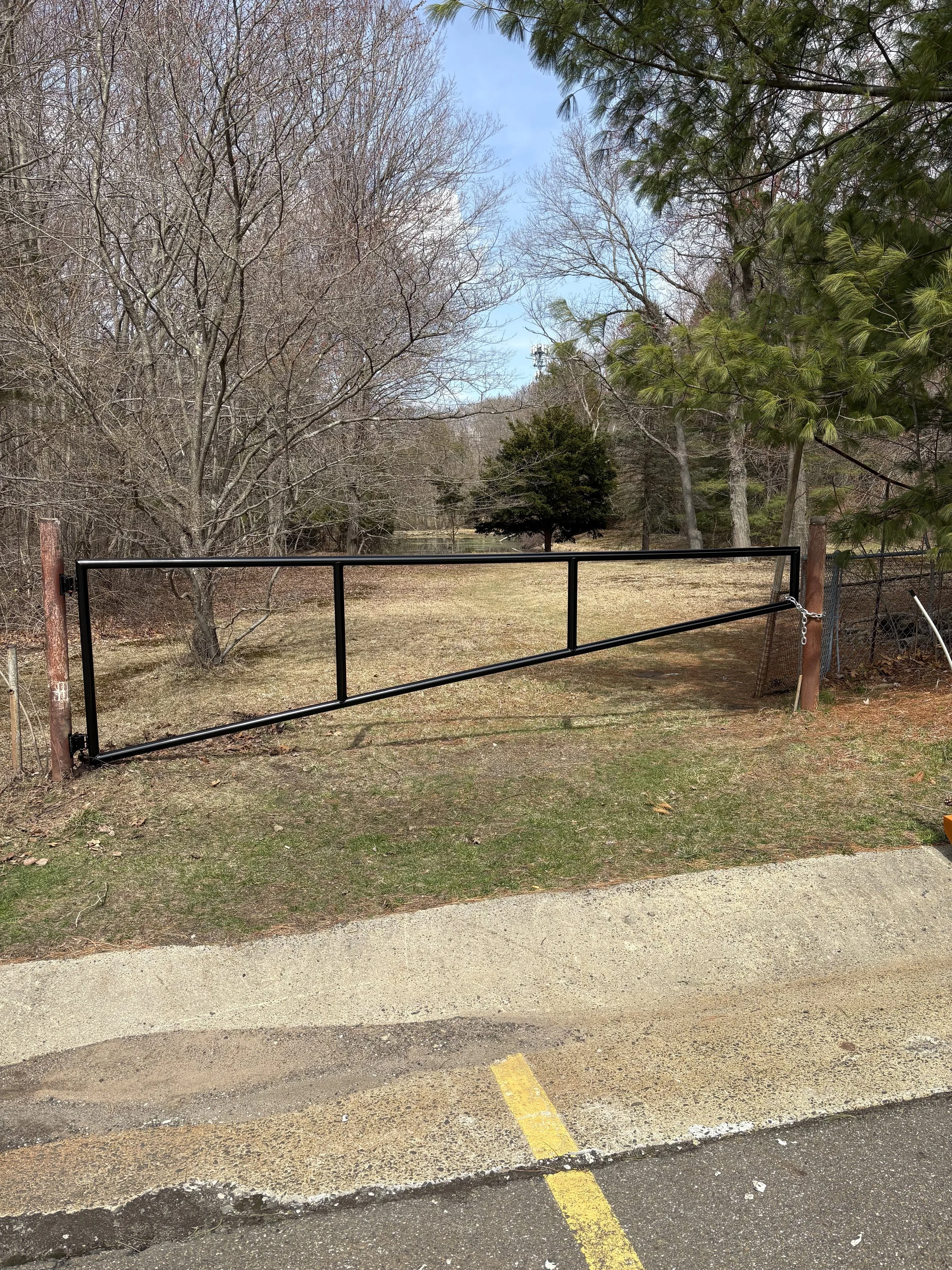 A black metal gate across a grassy pathway in a suburban area, with a paved parking lot in the foreground and leafless trees in the background.