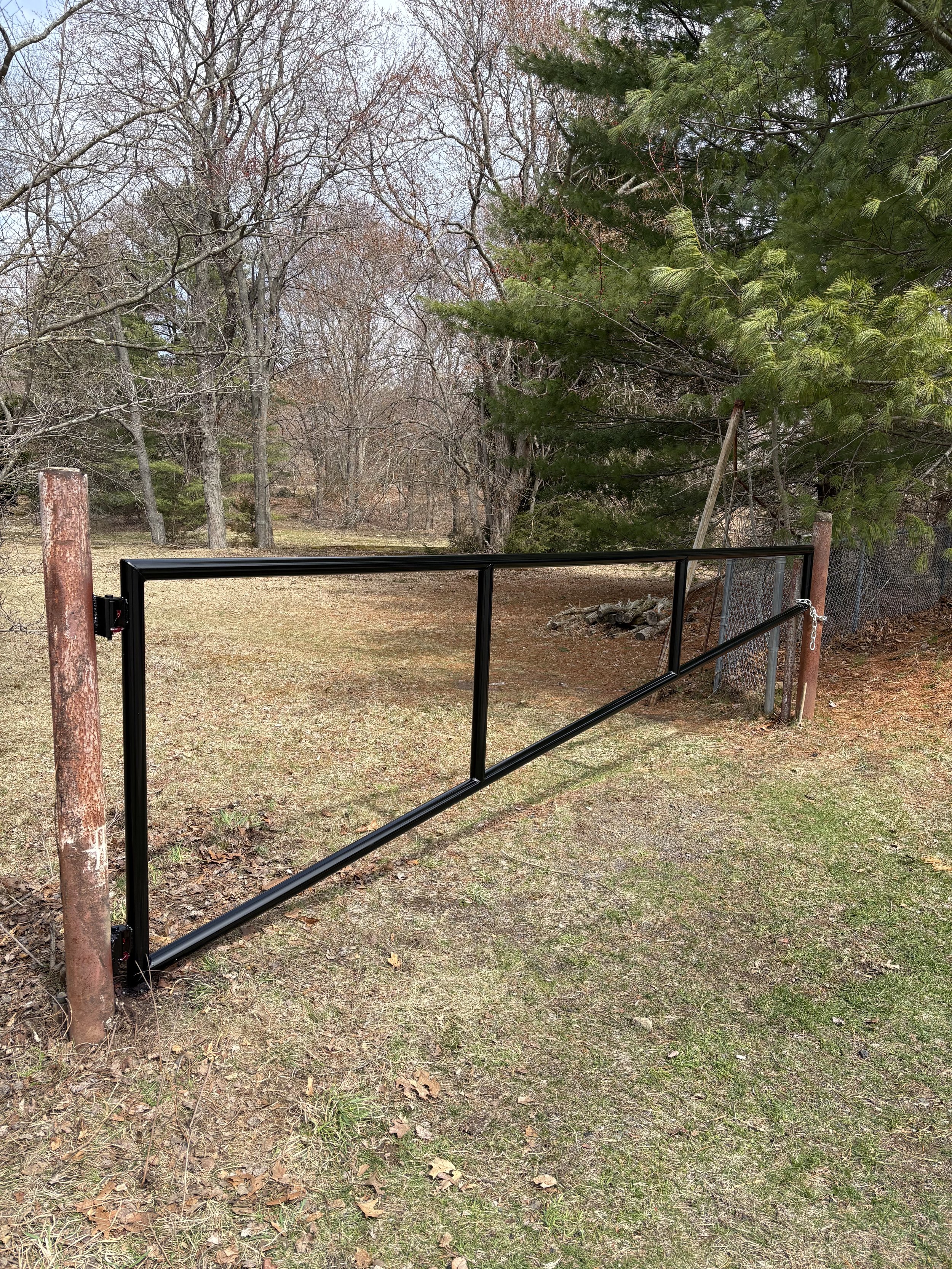 A metal gate with a chain and lock, installed between two weathered wooden posts, in a backyard with grass and trees.