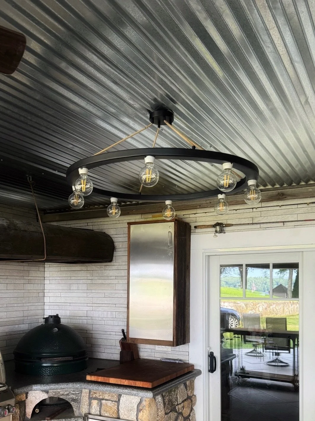 Indoor patio with decorative black circular chandelier featuring eight exposed light bulbs, adjacent to sliding glass door revealing an outdoor landscape with grass, trees, and a parked vehicle.