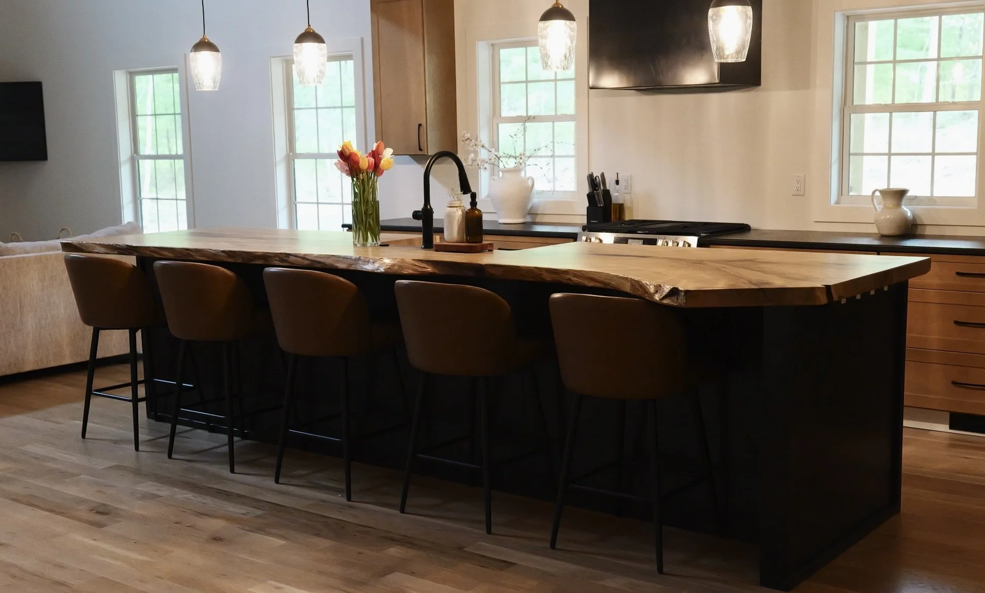 Modern kitchen with a large wooden island, six brown bar stools, pendant lights, black sink faucet, vase of tulips, and windows letting in natural light.