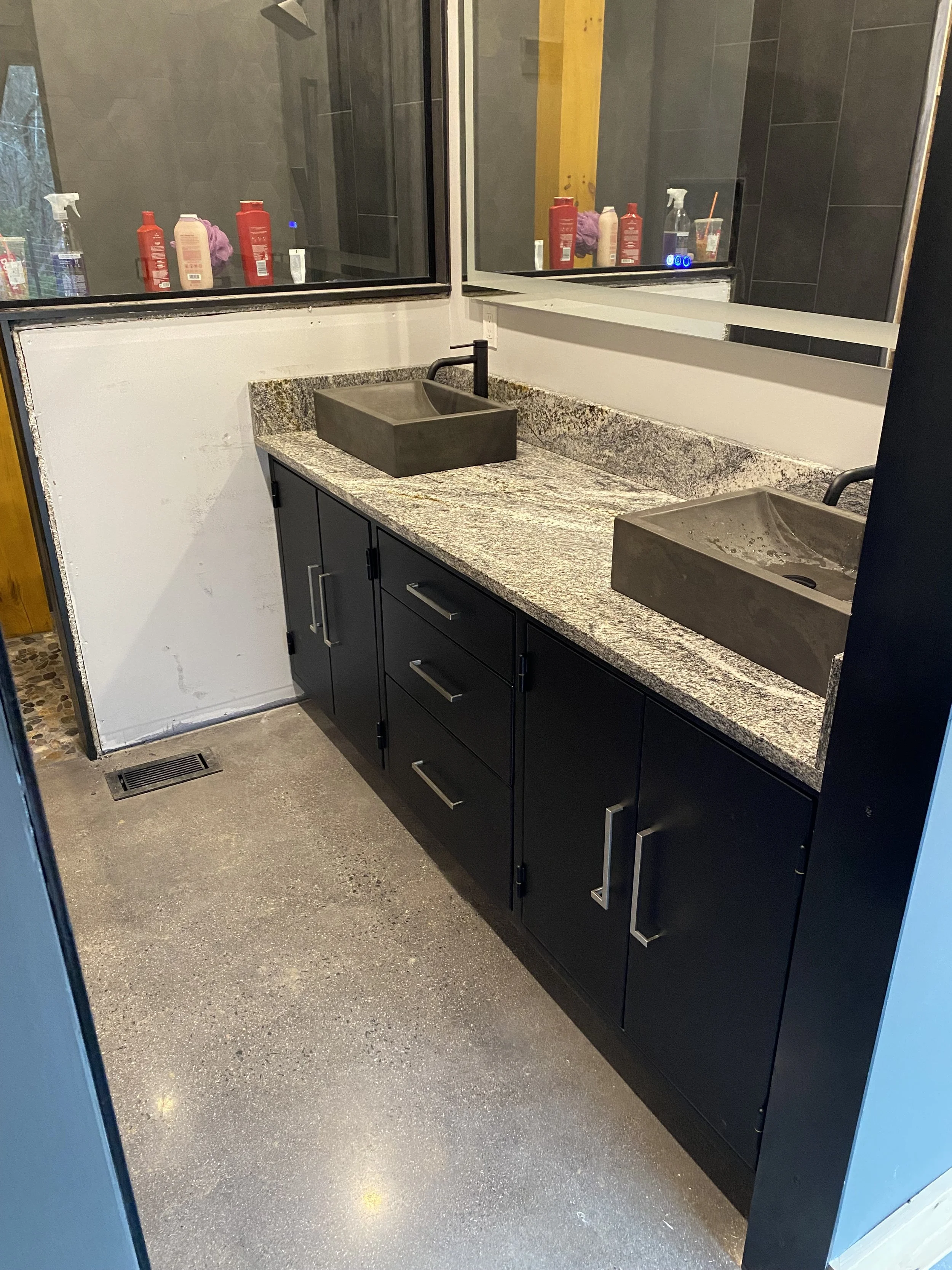 Double bathroom sink vanity with stone countertop, dark cabinets, and two rectangular vessel sinks with black faucets, mirror above, and cleaning supplies on upper shelf.