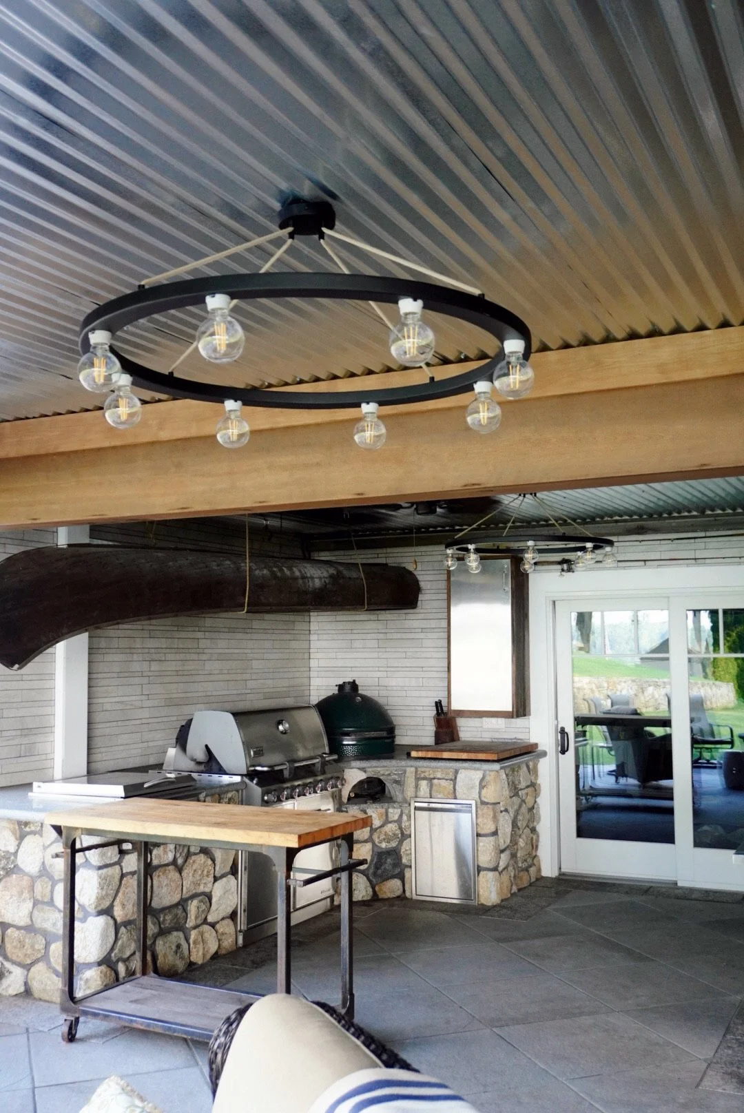 Outdoor kitchen with grill, stone counter, and large sliding glass door, featuring circular hanging light fixtures on a patio.