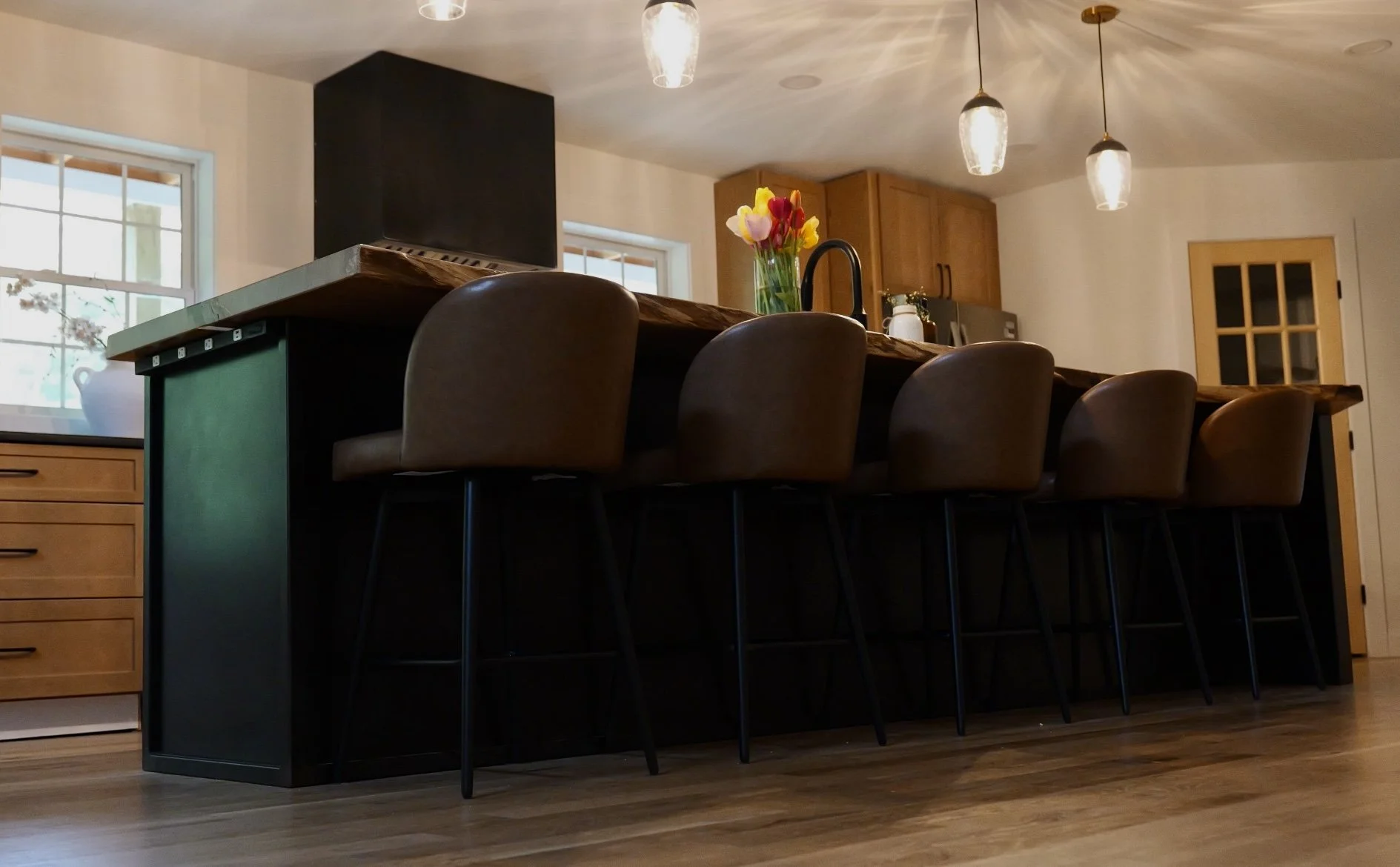 Kitchen with bar counter, brown chairs, vase with colorful tulips, windows, and modern light fixtures.