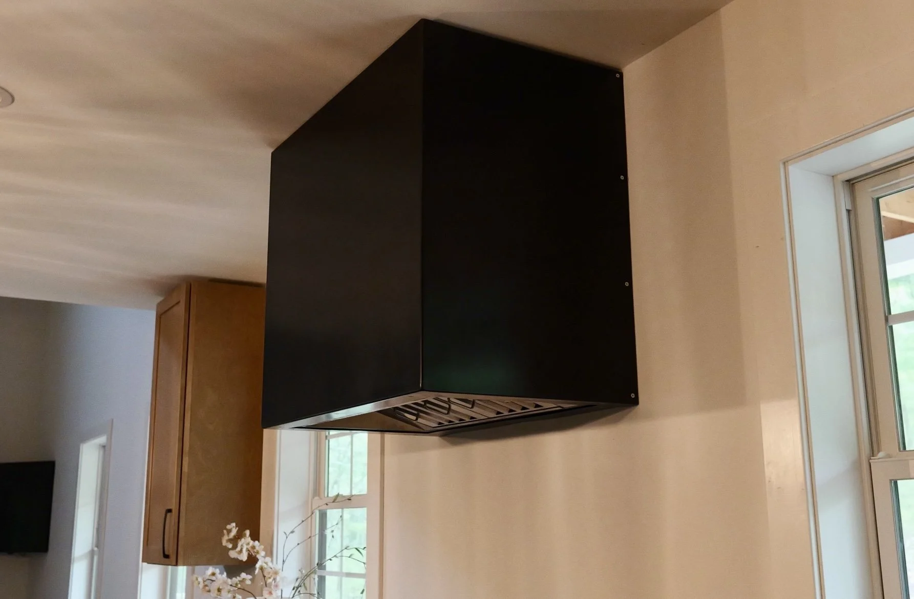 Black kitchen range hood above a stove in a kitchen with beige walls and large windows.