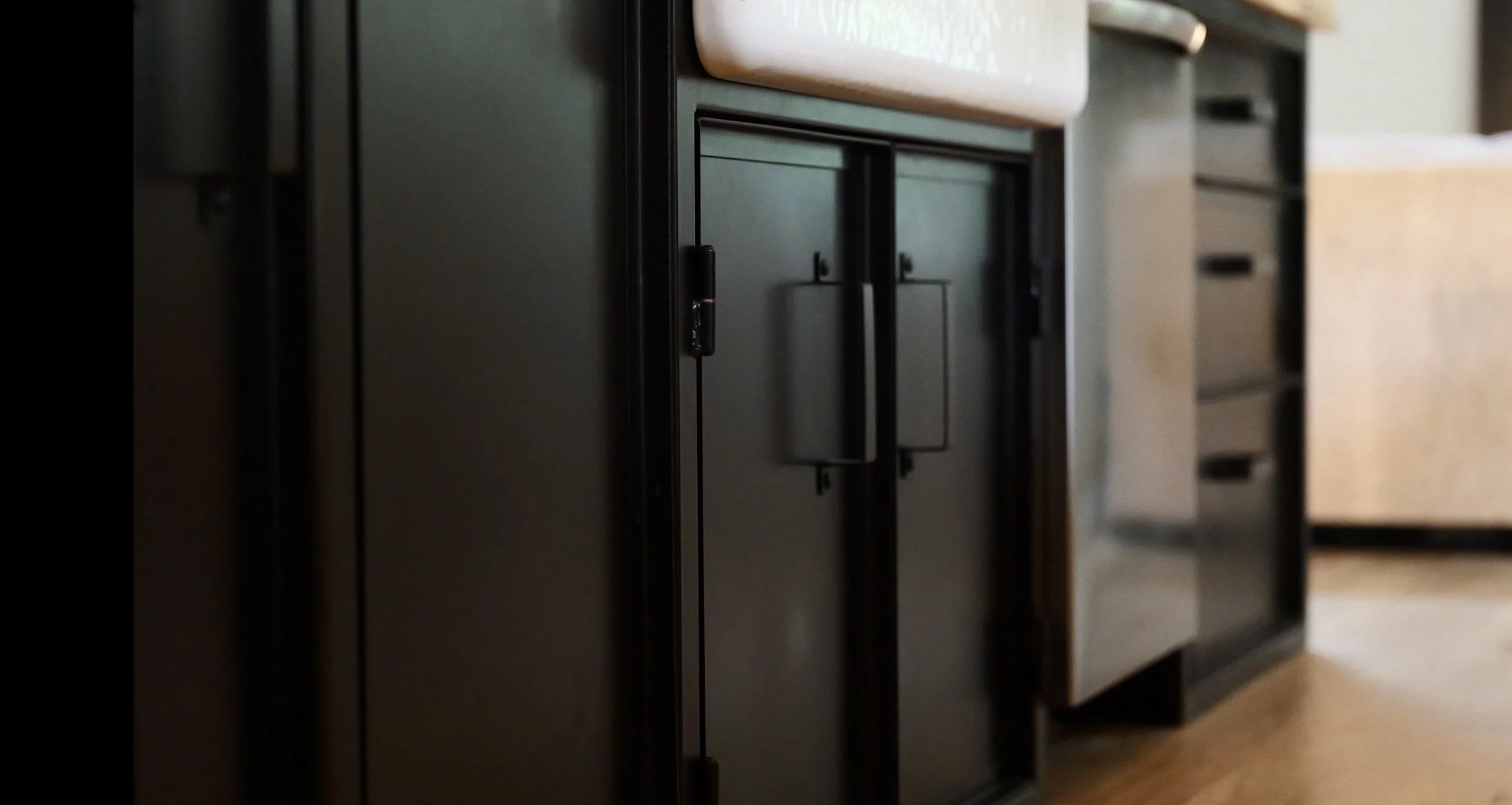 Close-up of black refrigerator with a handle and door, in a kitchen with beige and black cabinets and wooden floor.
