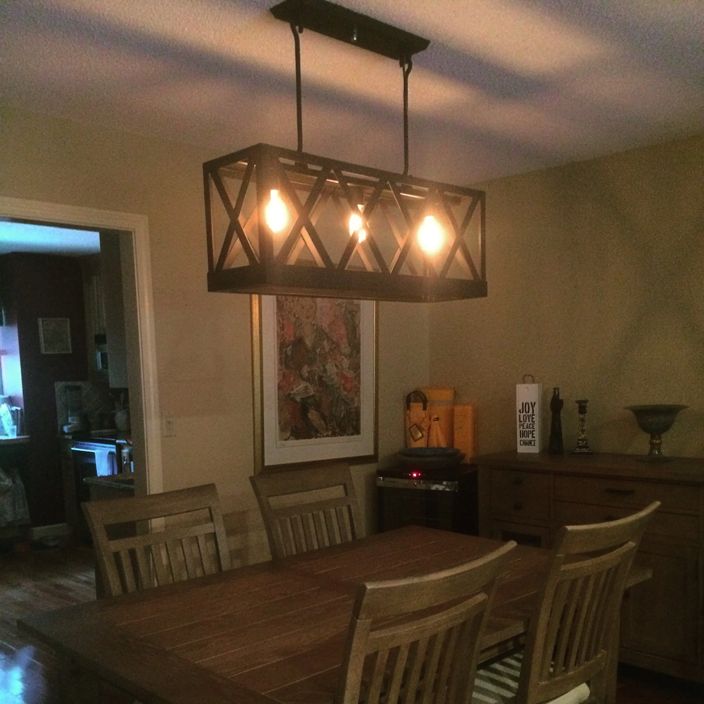 A dining room with a rectangular wooden table, surrounded by six wooden chairs, a chandelier with exposed bulbs hanging overhead, a dresser with decorative items including a sign with words like JOY, LOVE, PEACE, HOPE, and a framed artwork on the wal