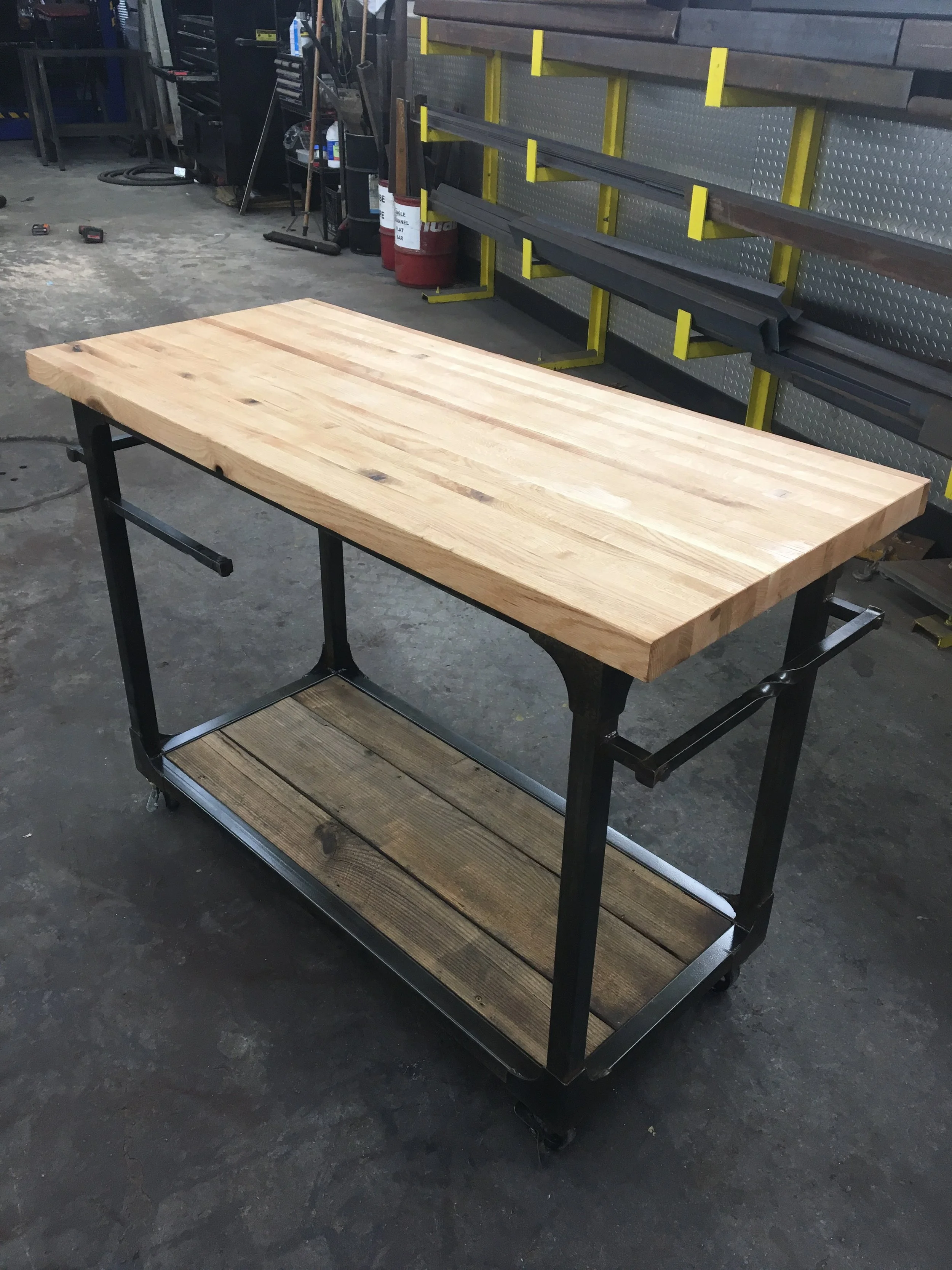 A workshop table with a wooden top and a lower shelf made of wood planks, supported by a metal frame on wheels, located in a workshop with tools and shelves in the background.
