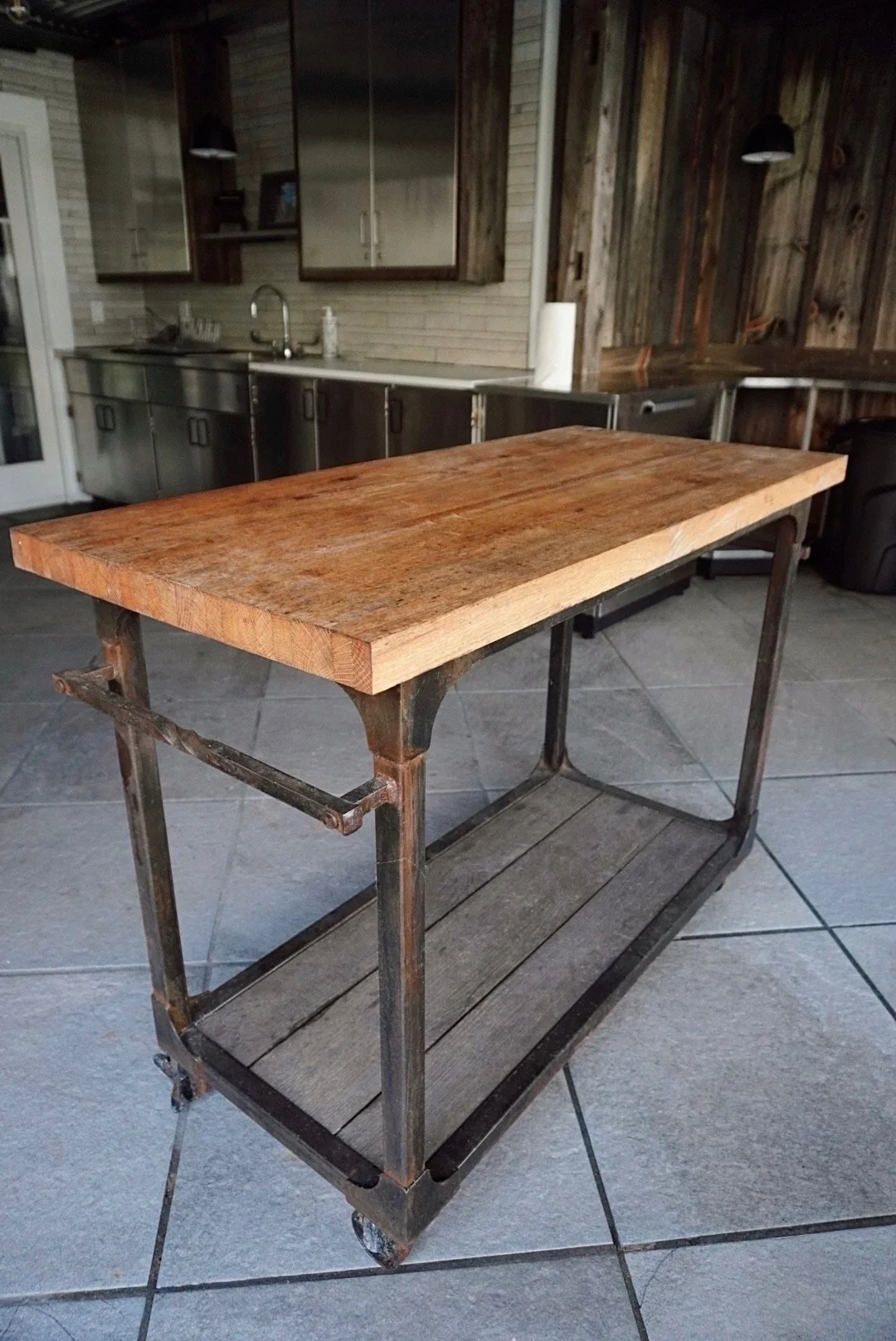 A rustic kitchen island with a wooden top and metal frame with wheels, in a kitchen with tiled floor and wooden wall decor.