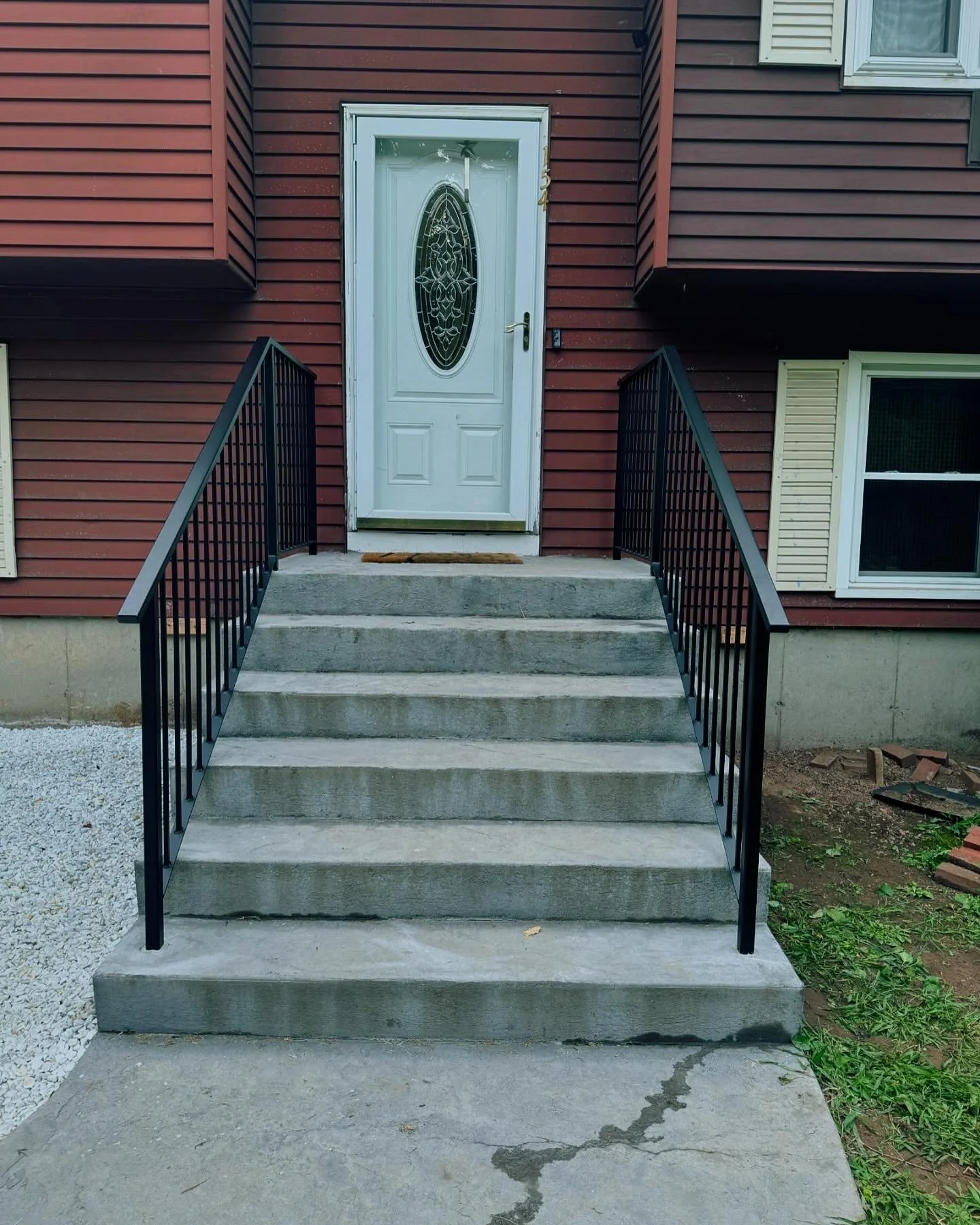 Concrete steps with black metal railing leading up to a white front door with an oval decorative glass panel, outside a red house with beige shutters.
