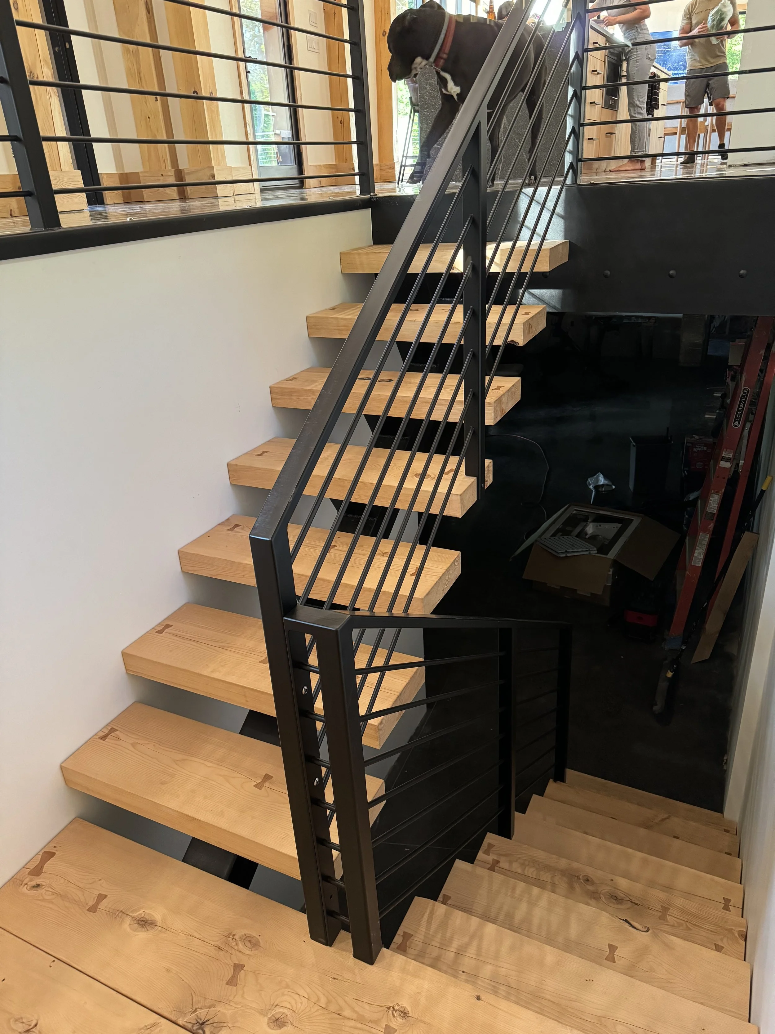 Interior staircase with wooden steps and black metal railing, leading to an upper level with people standing.