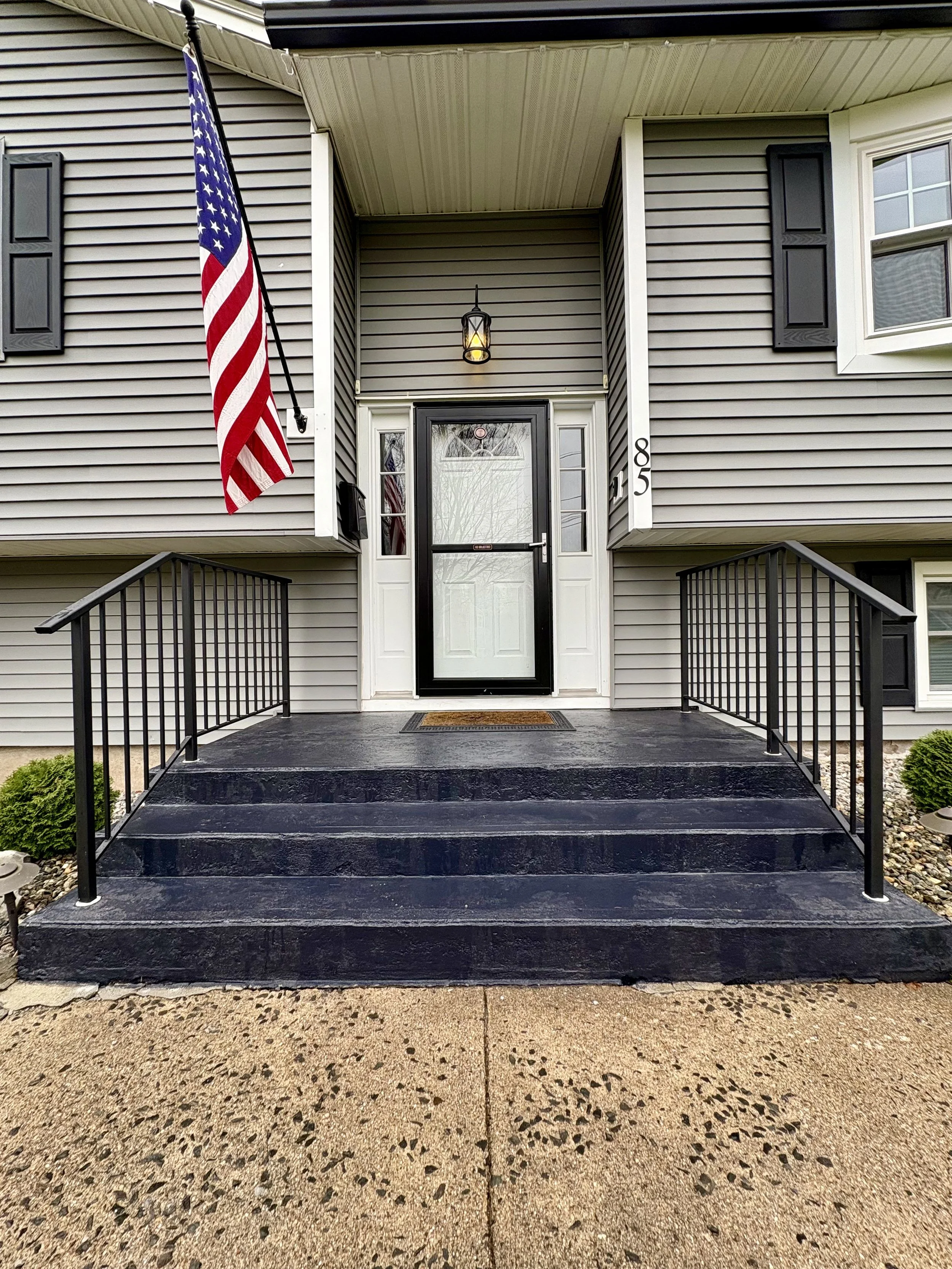Front porch of a house with a black painted deck, black metal railing, a white door with glass panels, an American flag hanging on the left, a hanging lantern light above the door, gray vinyl siding, and a small green shrub on each side of the steps.
