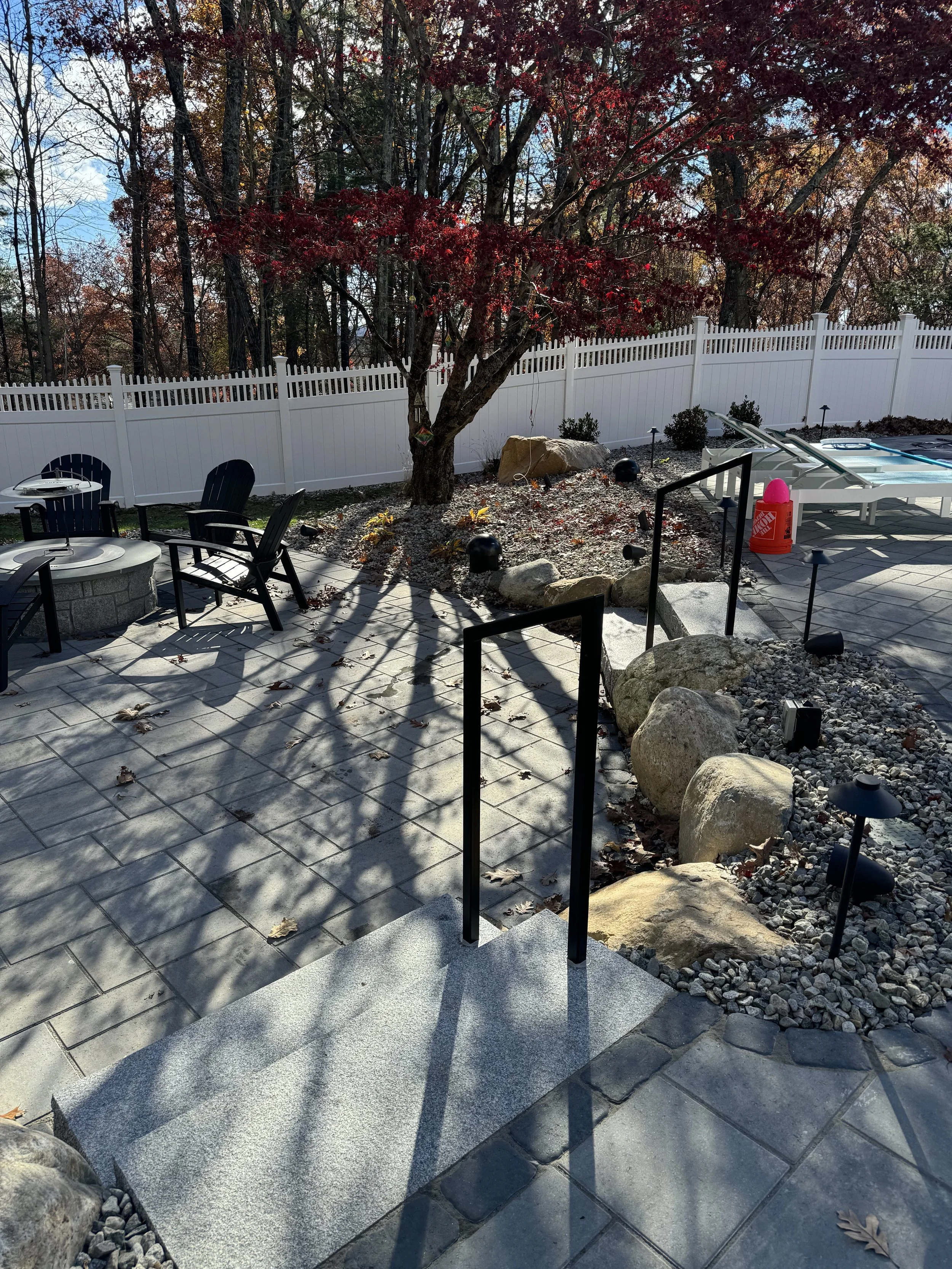 A backyard patio with a white picket fence, red leafed tree, outdoor chairs around a fire pit, and stepping stones leading to a swimming pool, with fall leaves scattered on the ground.