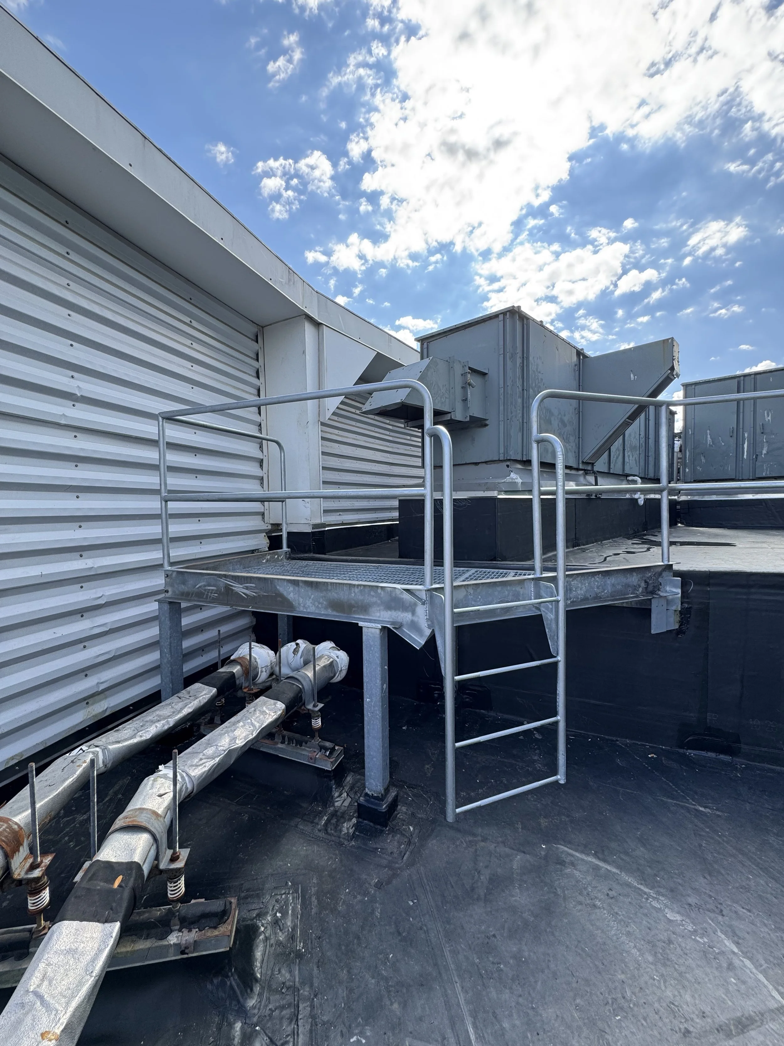 Rooftop HVAC system with metal ducts, pipes, and safety railing against a blue sky with clouds.